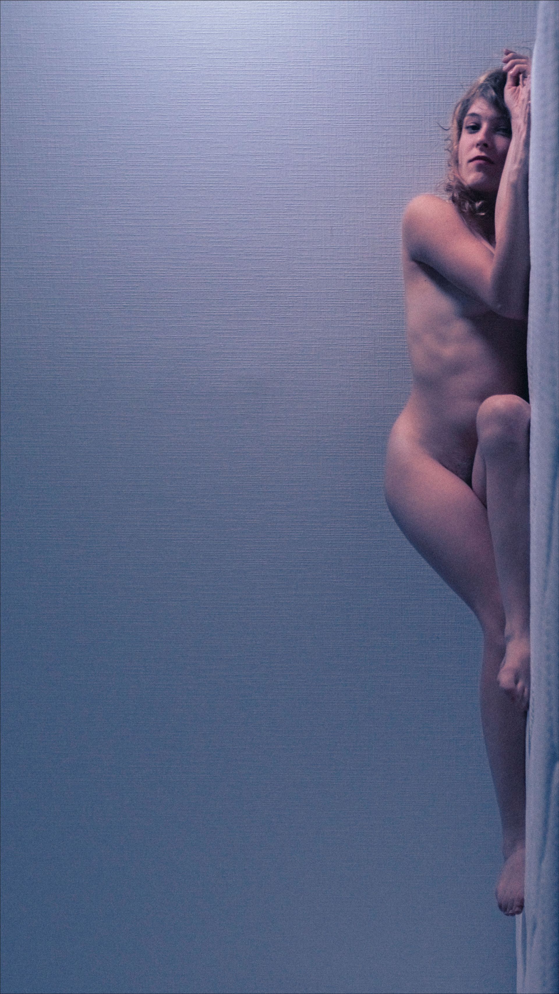 Nude person leans against a textured, light-colored wall with blue background.