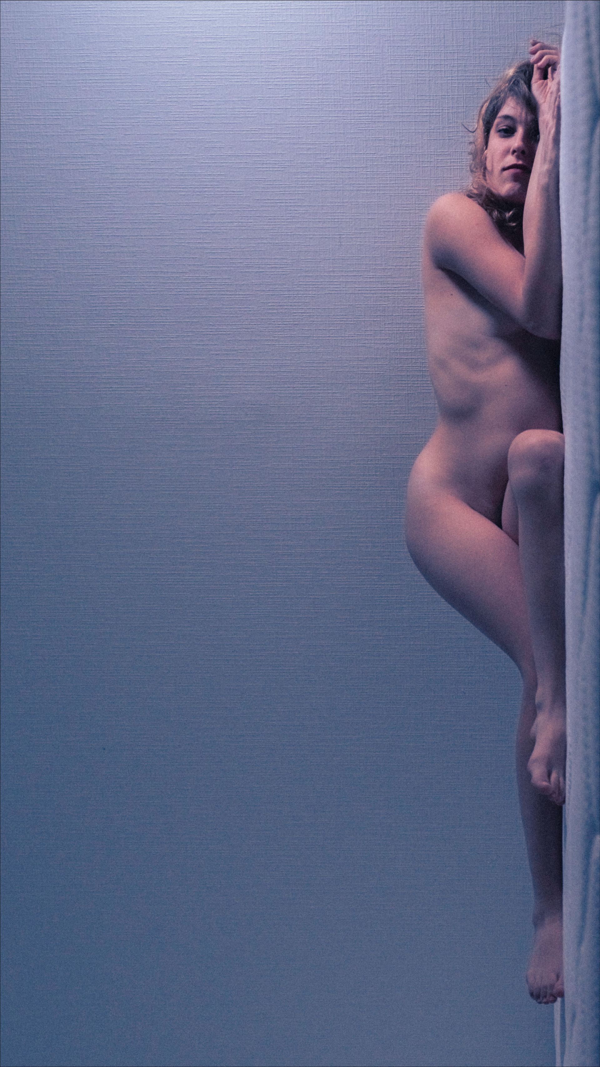 Nude person leaning against a textured wall, arms over head, light skin, blue tones.
