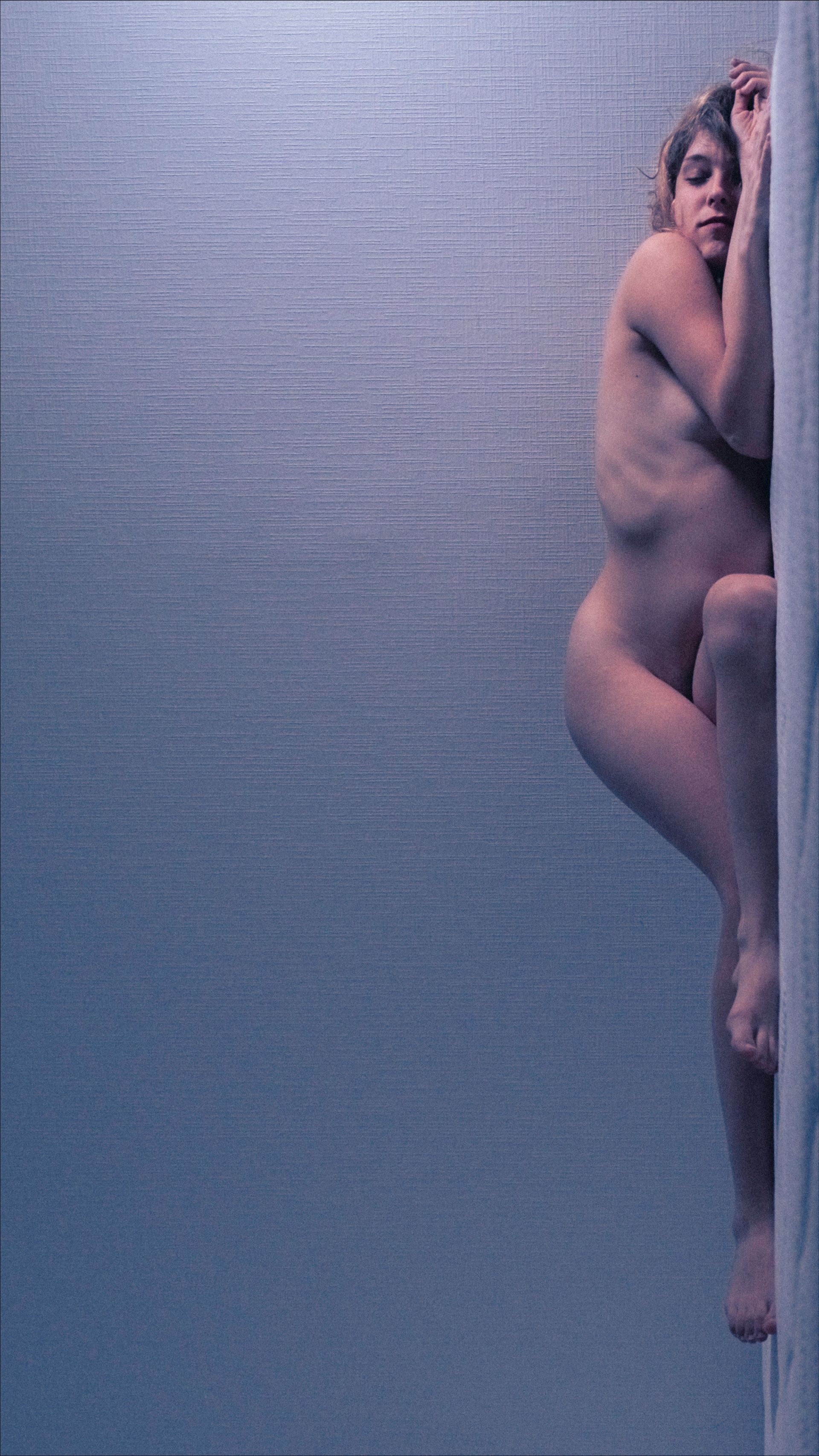 Nude person leans against a light blue wall. Eyes are closed, head resting.