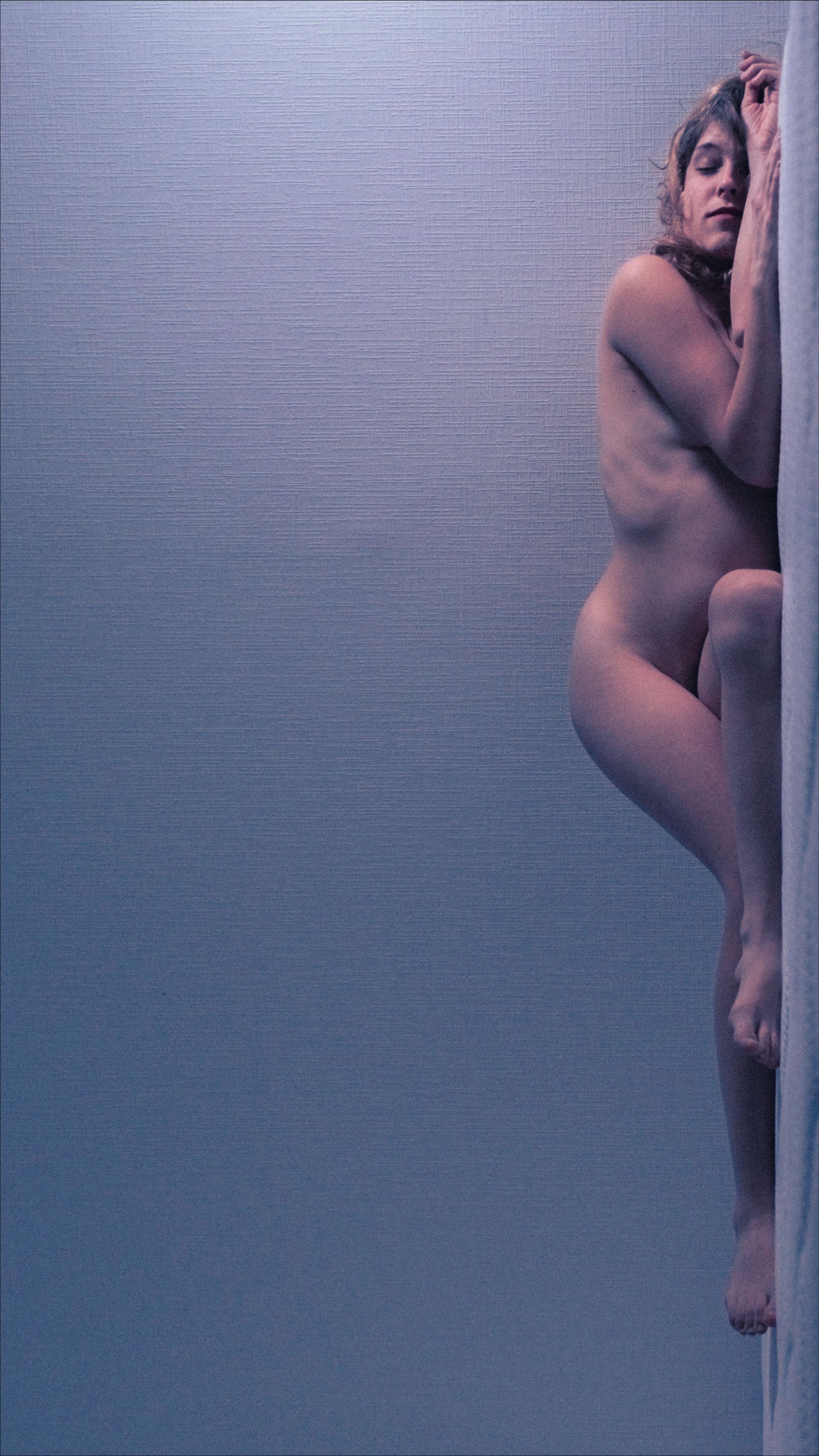 Nude person leaning against a textured wall, arms wrapped around a column.