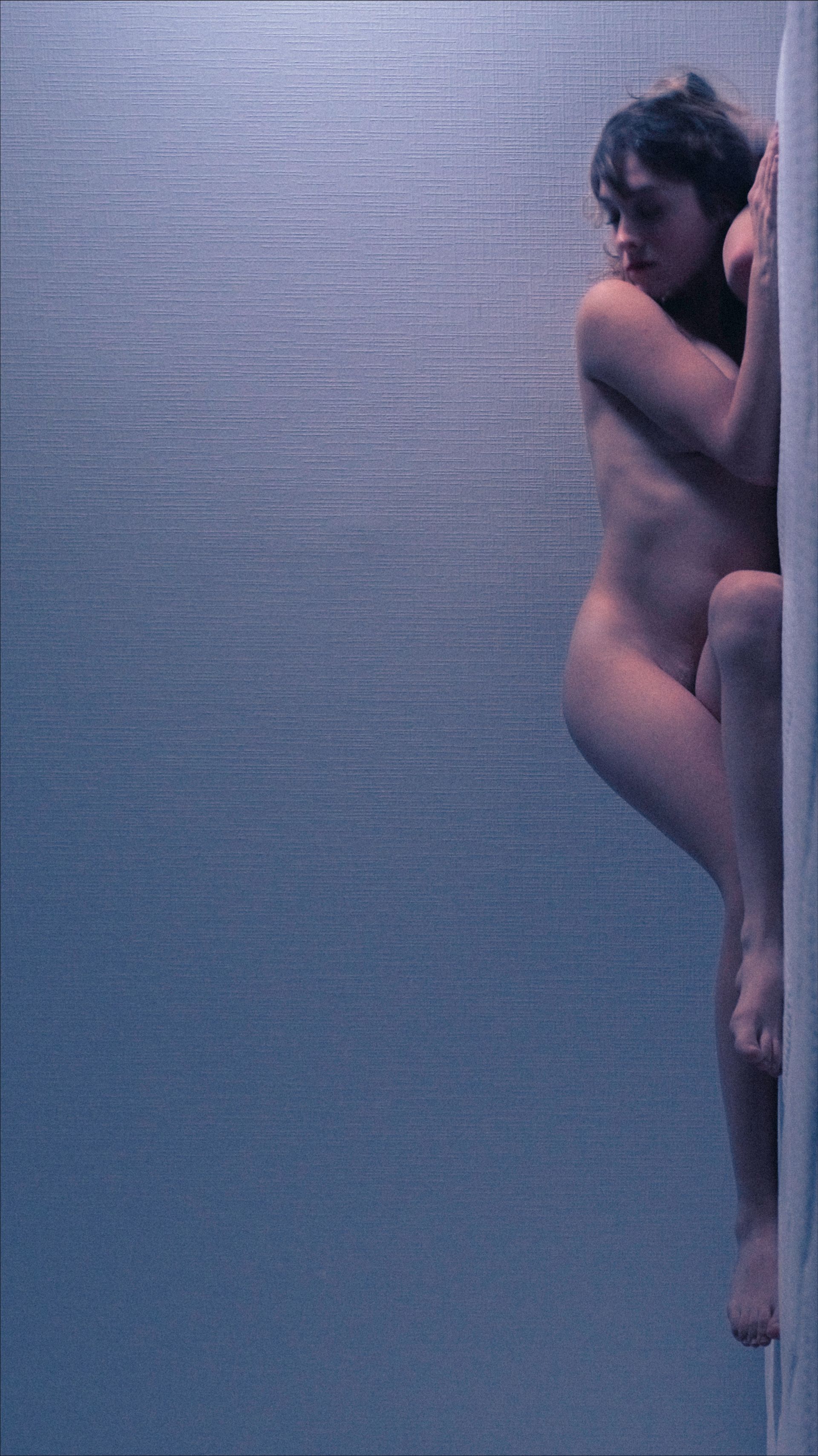 Nude person leaning against a light wall, in a blue-tinted space, arms around head.