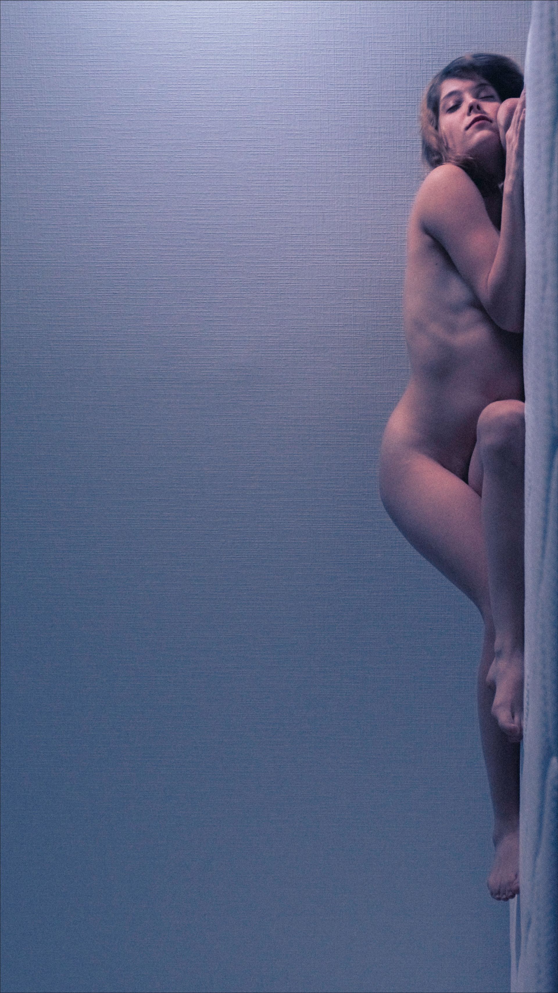 Nude person clinging to a textured, light-colored wall with blueish tones.