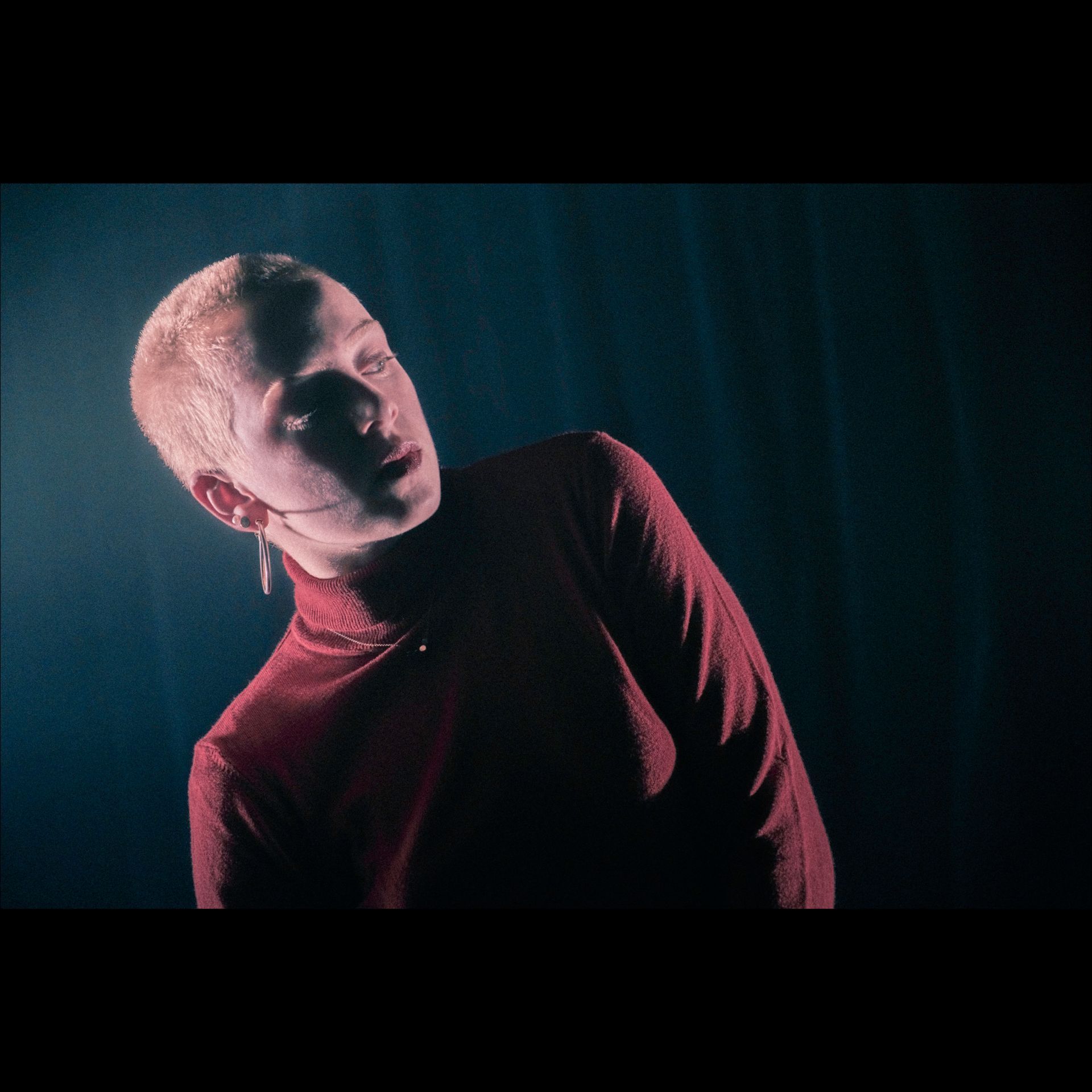 Person with short, bleached hair in a red turtleneck, looking upward with a serious expression, dark background.