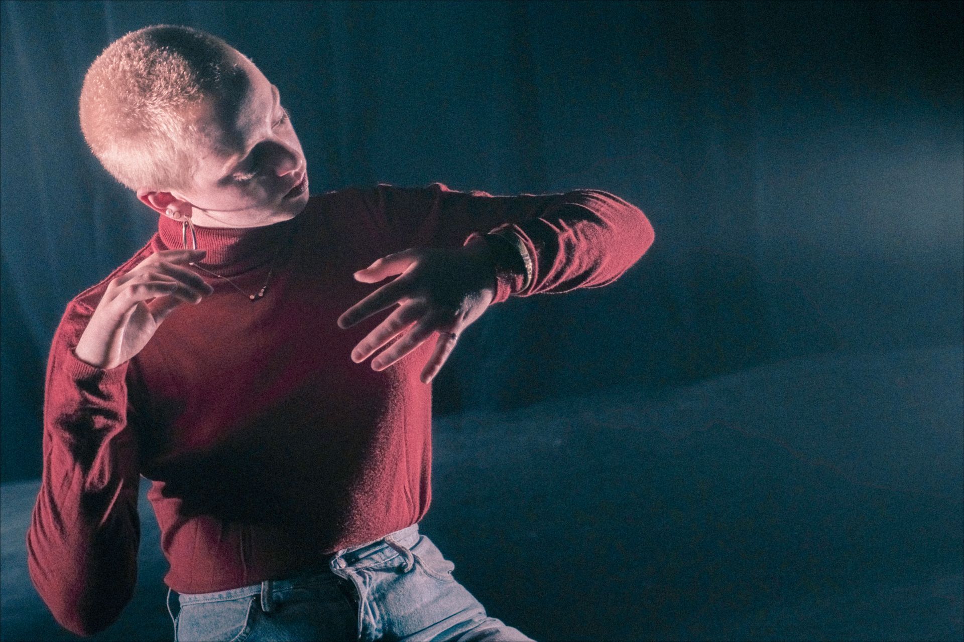 Person in red turtleneck and jeans, arms outstretched, dancing against a dark background.