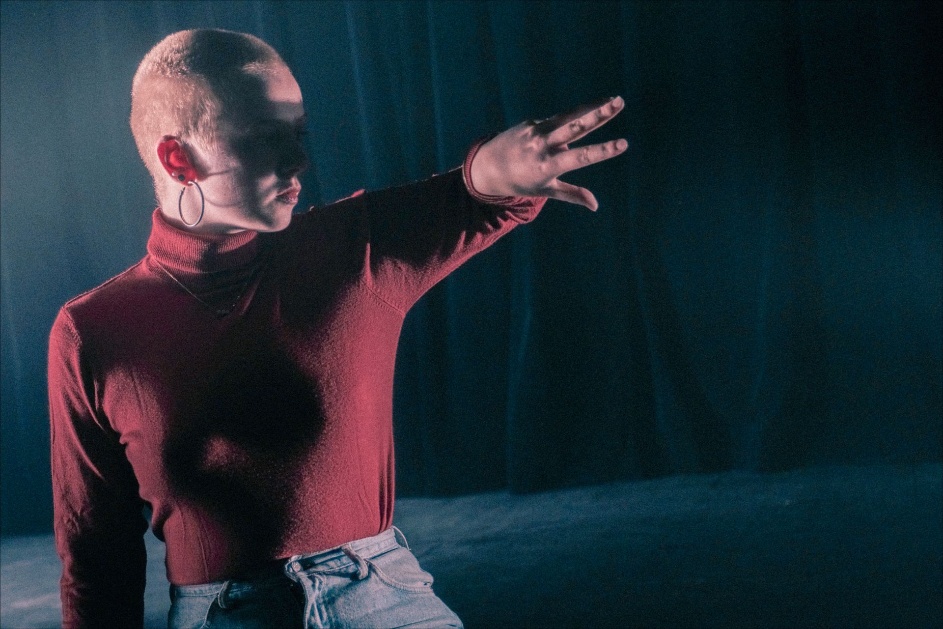 Person with shaved head in red turtleneck, hand extended, dark setting.