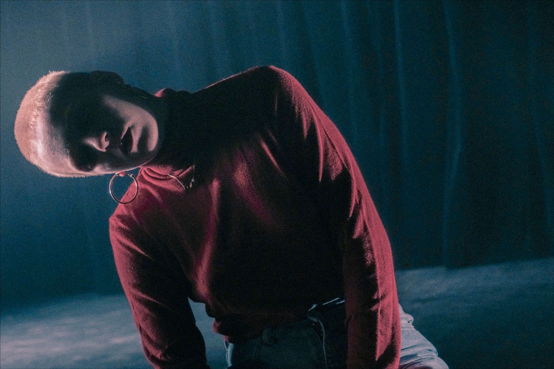 Person with shaved head in red shirt, tilted head, dark background, possibly rain.