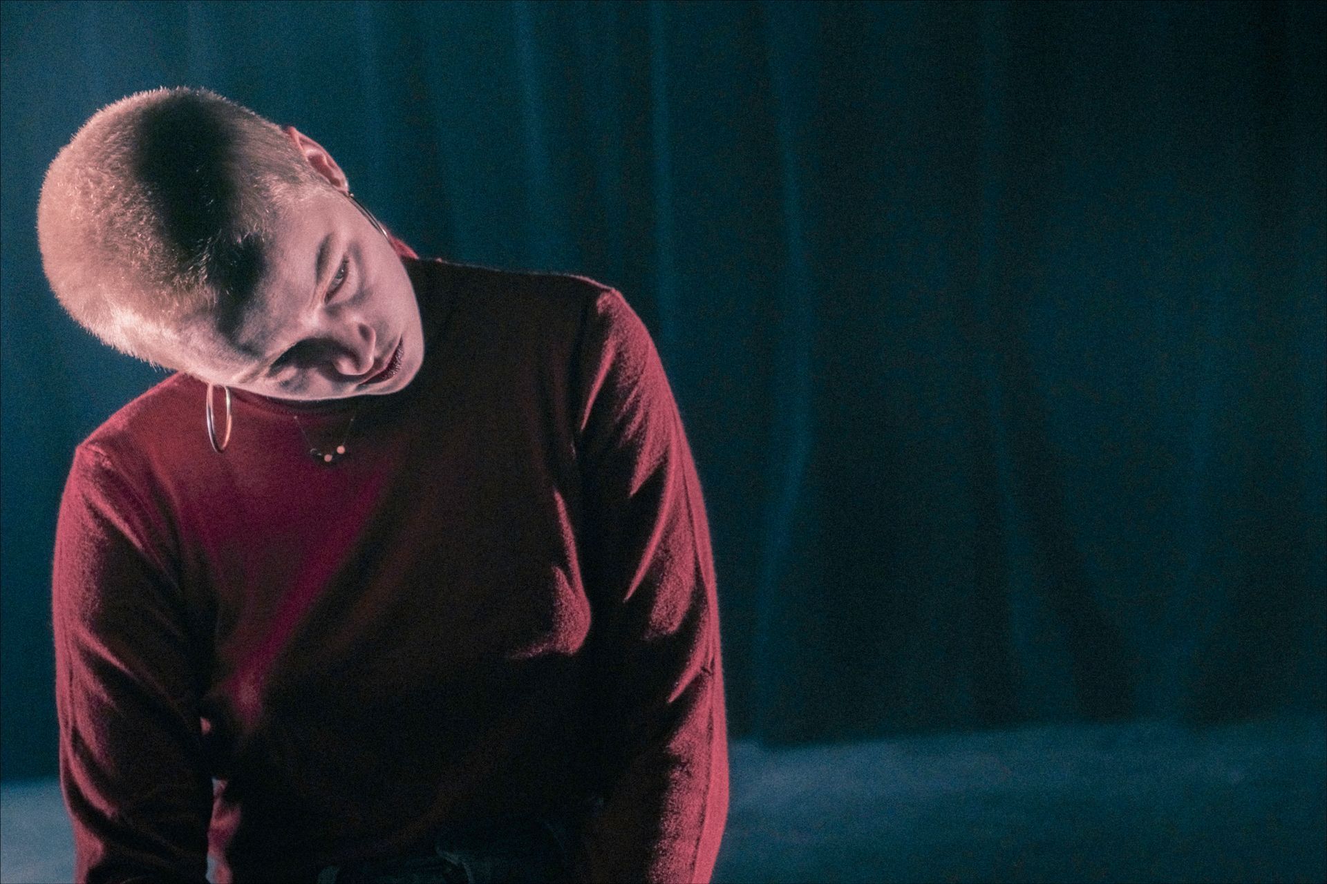 Person with shaved head wearing a maroon shirt, tilted head down. Dim, dark setting.