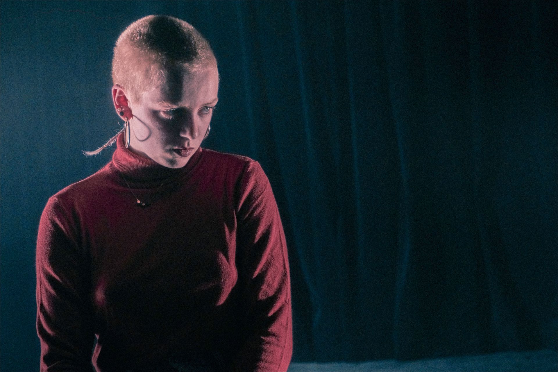 Person with shaved head in red turtleneck, looking down. Dark backdrop, lit from above.