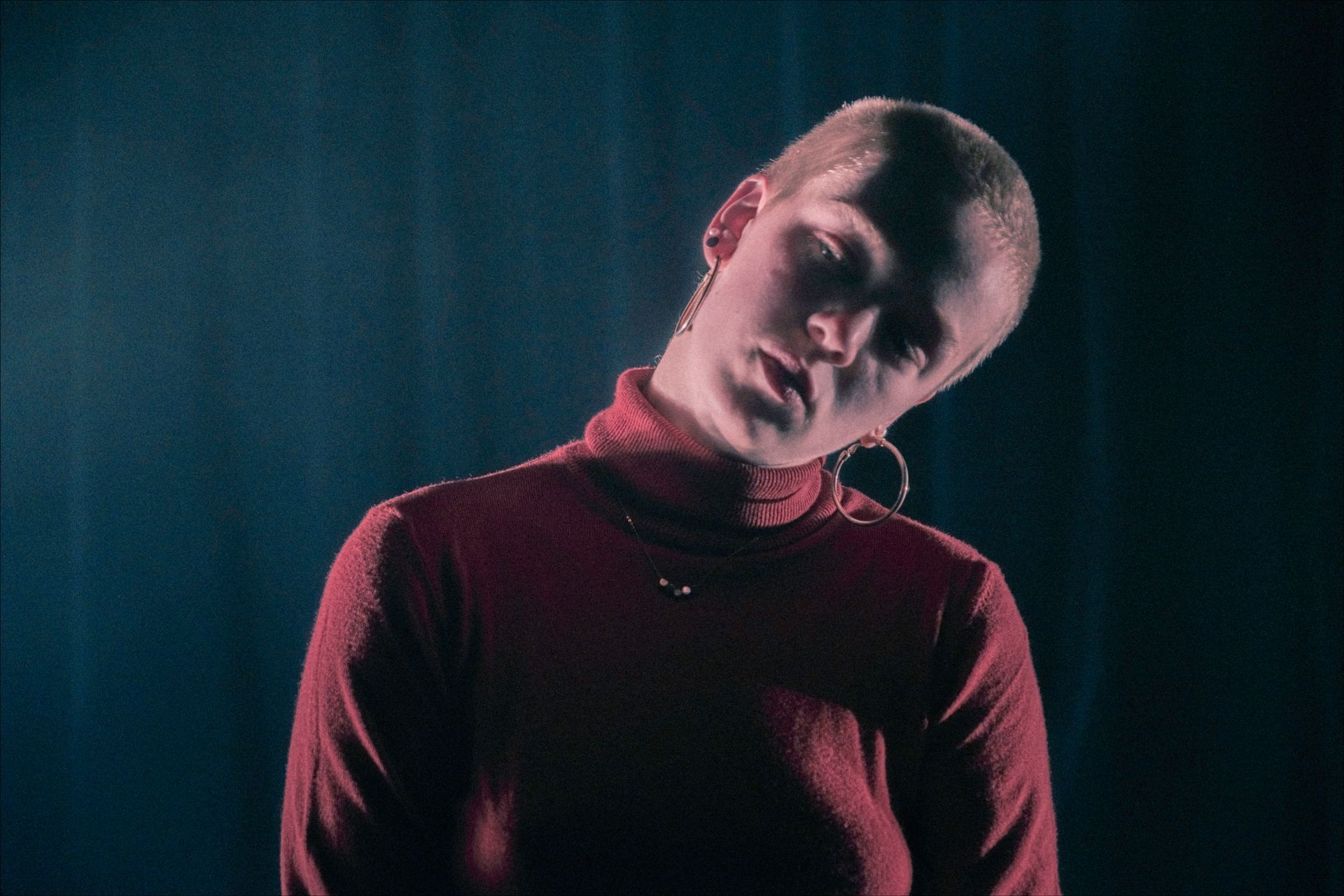 Woman with shaved head in red turtleneck, tilting head against a dark blue background.