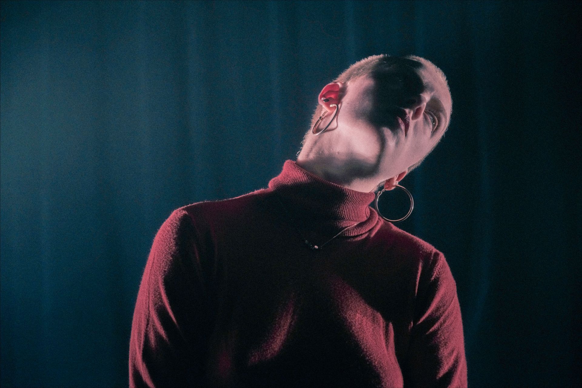 Person with shaved head wearing a red turtleneck, tilting head back. Dark background.