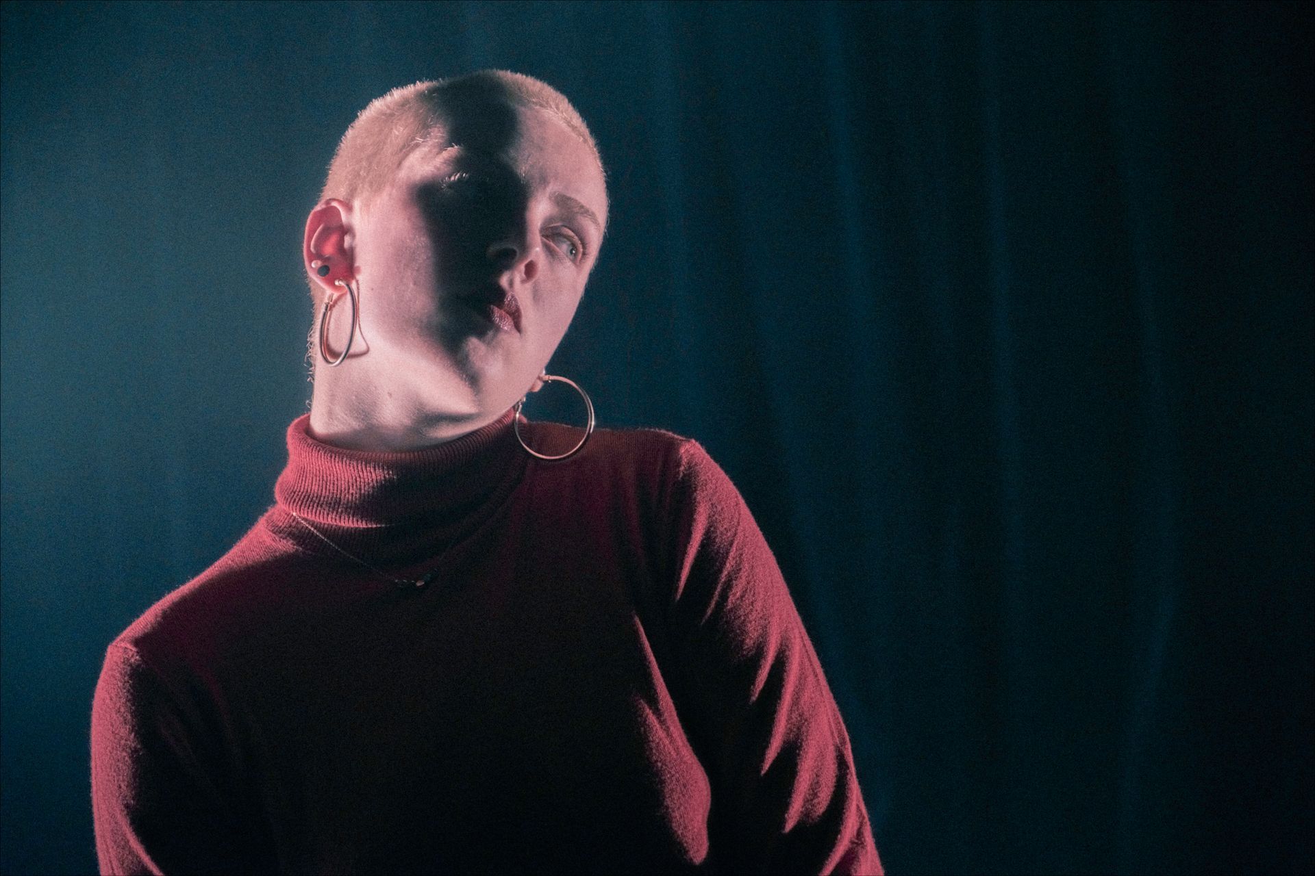 Person with shaved head and hoop earrings, wearing a red turtleneck, lit against a dark background.