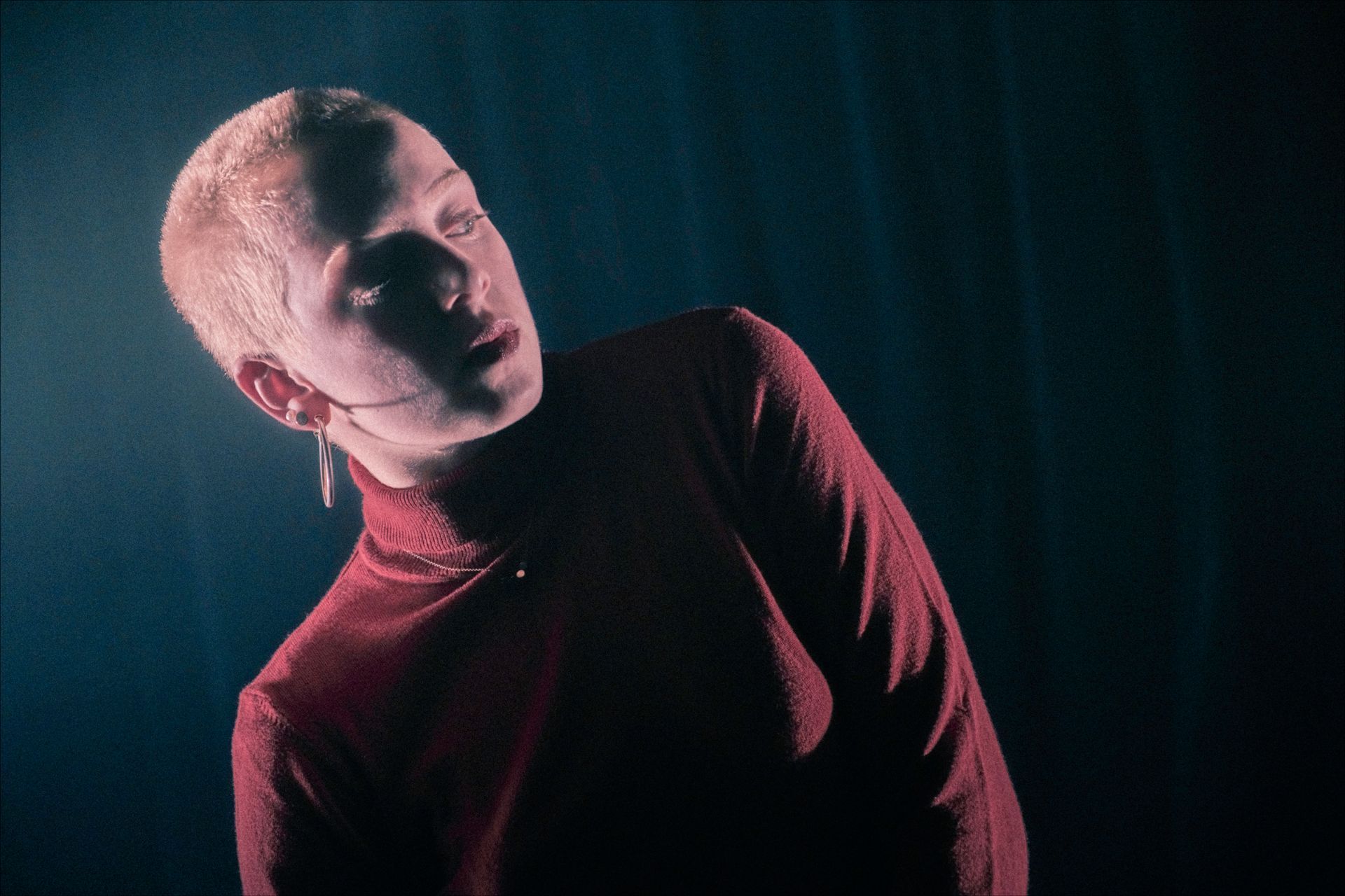 Person with short blond hair wearing a red turtleneck, looking upward with a slight smile, against a dark blue background.