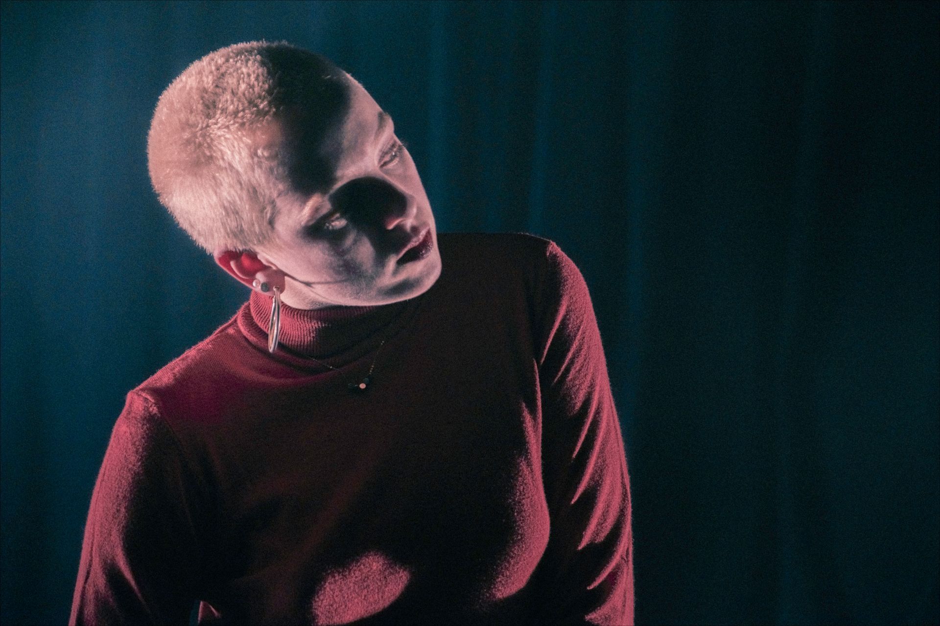 A person with short blonde hair looks upwards, wearing a red turtleneck, in a dark setting.