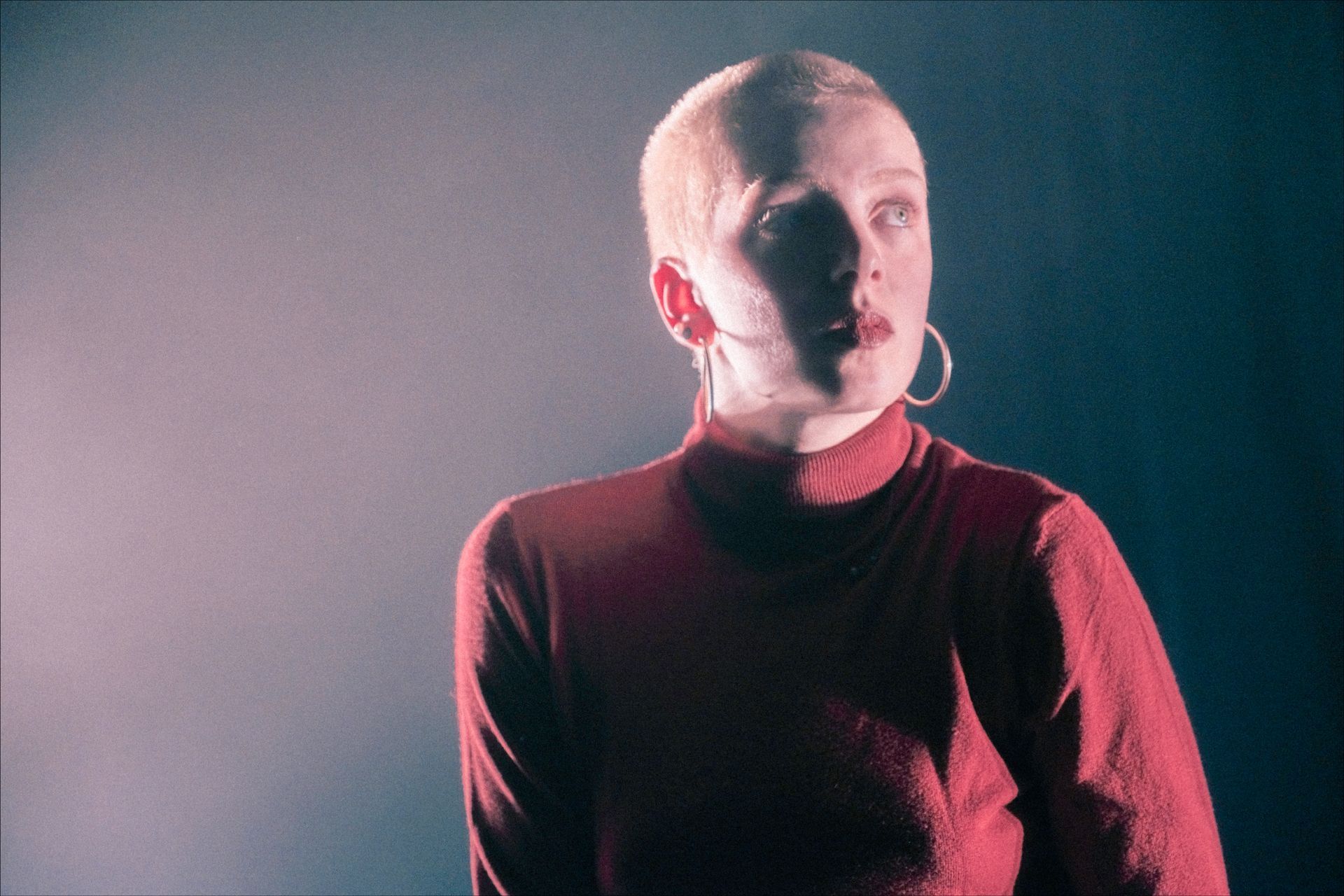 Woman with shaved head, wearing a red turtleneck, looking upward with a serious expression, dark backdrop.