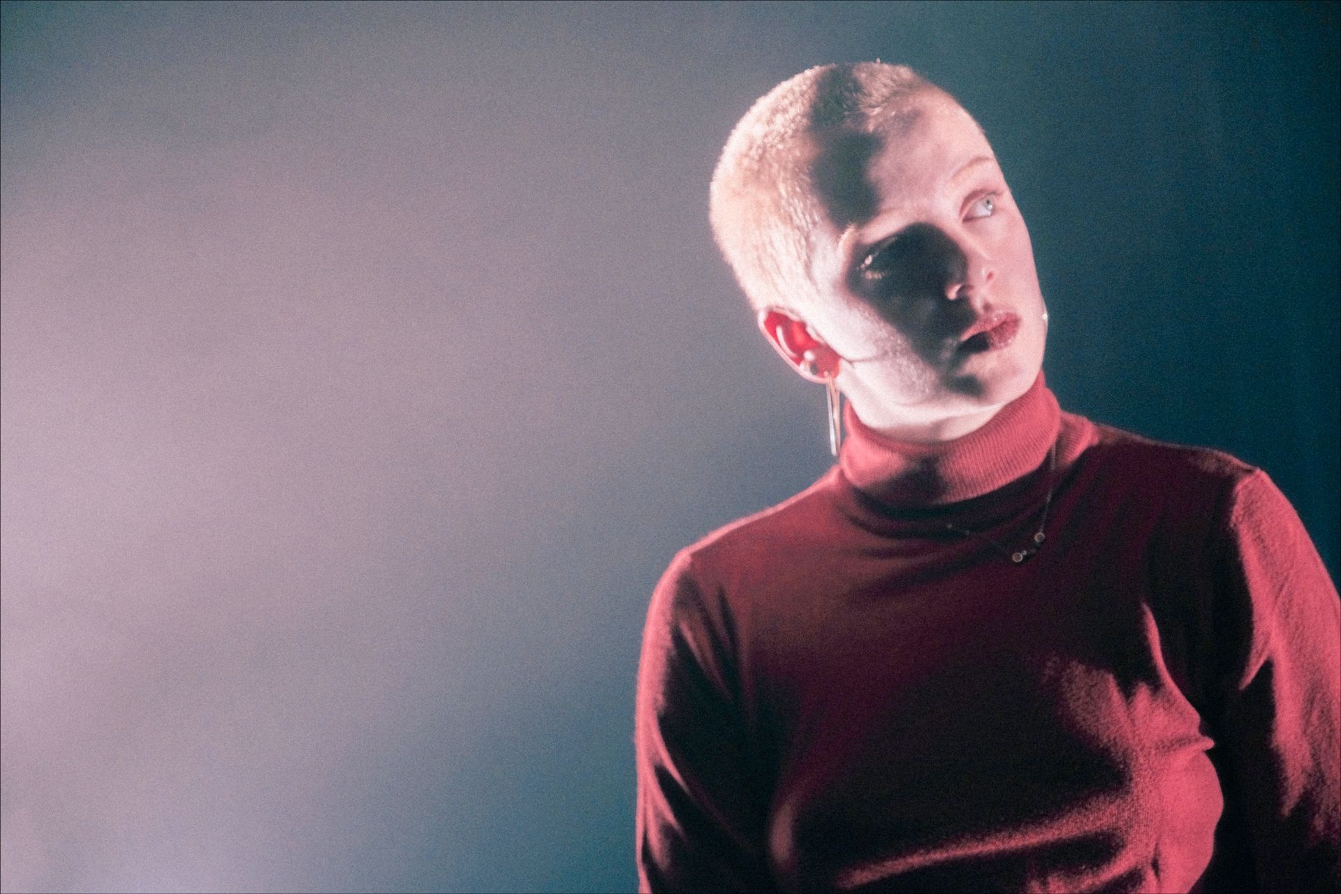 Person with shaved head in red turtleneck, looking upward with a slight smile in a dark, atmospheric setting.