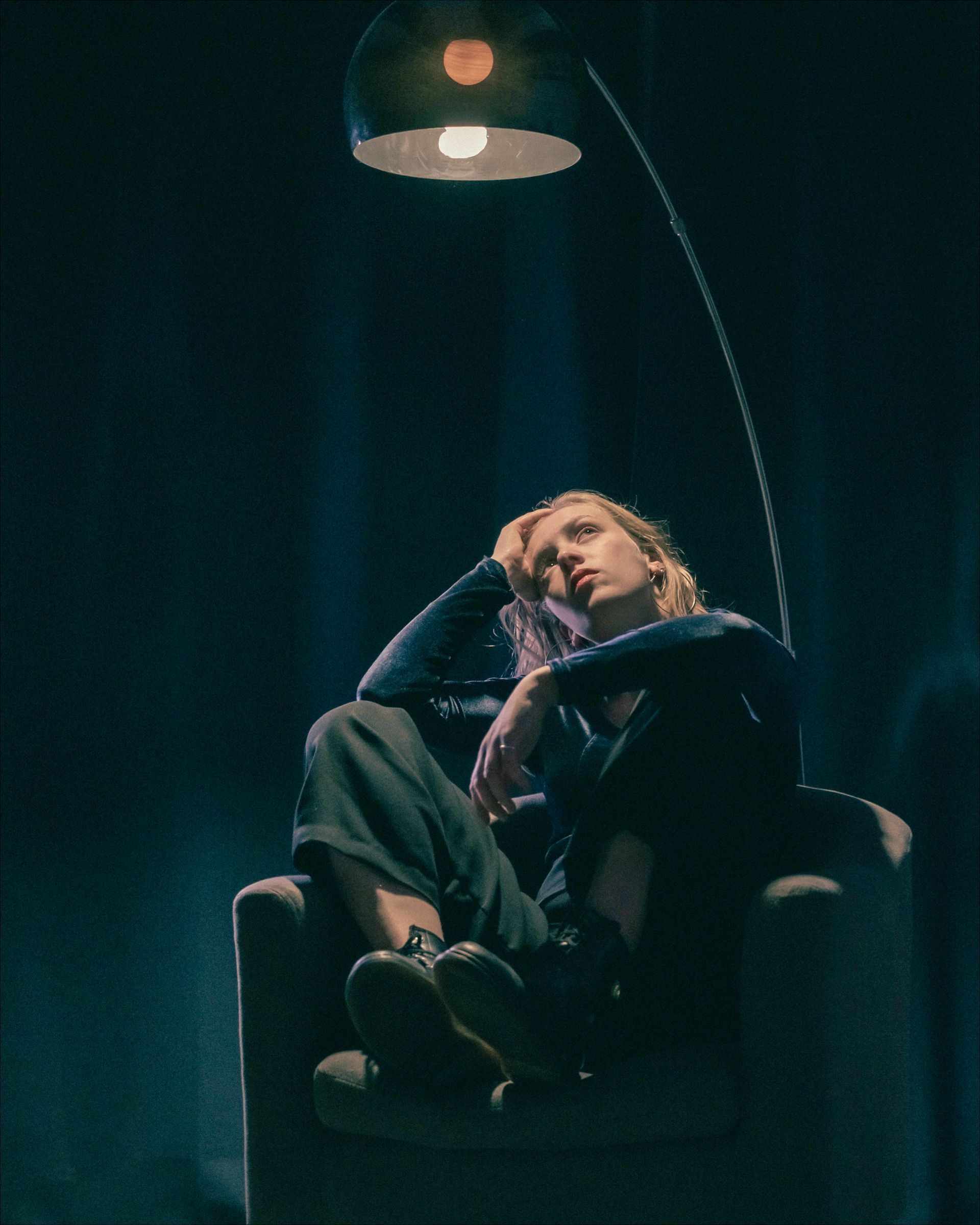 Person seated under a lamp, resting head on arm, dark clothing.