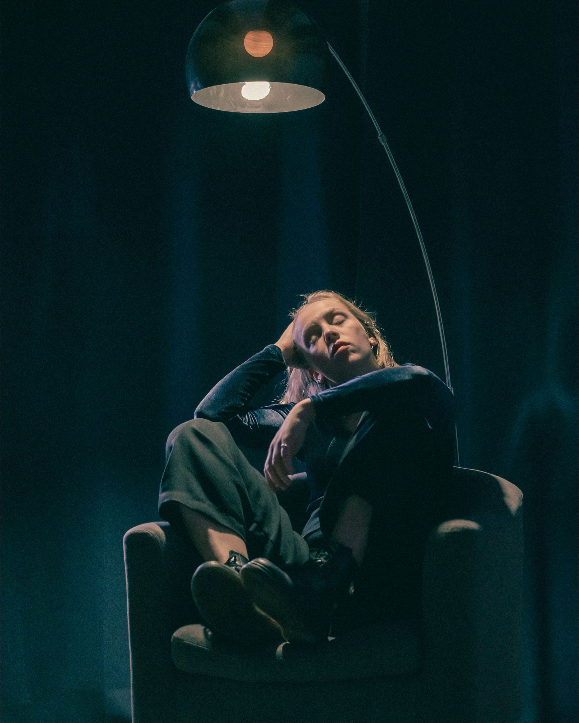 Person seated in armchair under a lit lamp, eyes closed, resting head on hand. Dark setting.