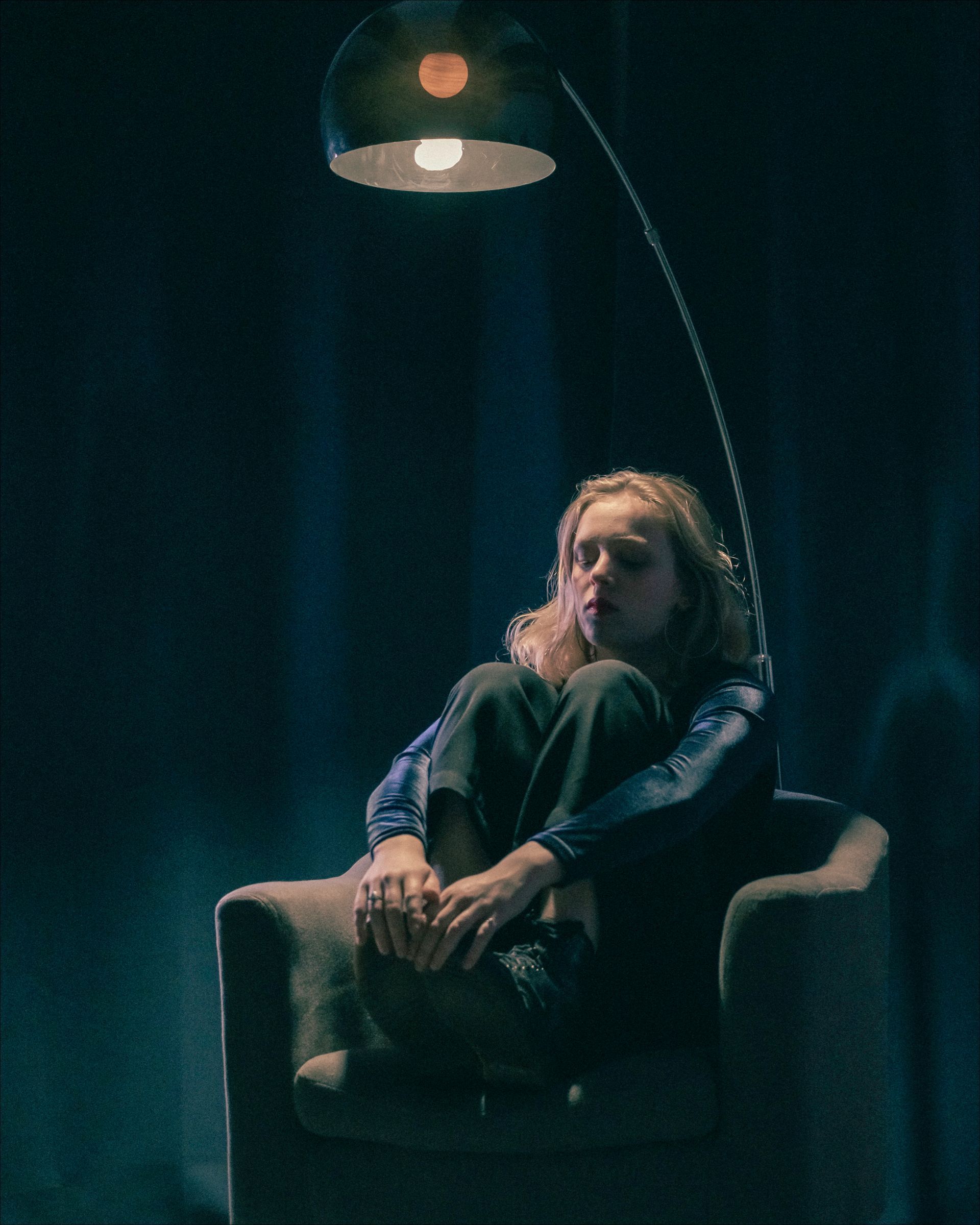 Person sits in an armchair under a curved lamp. Low light, moody atmosphere.