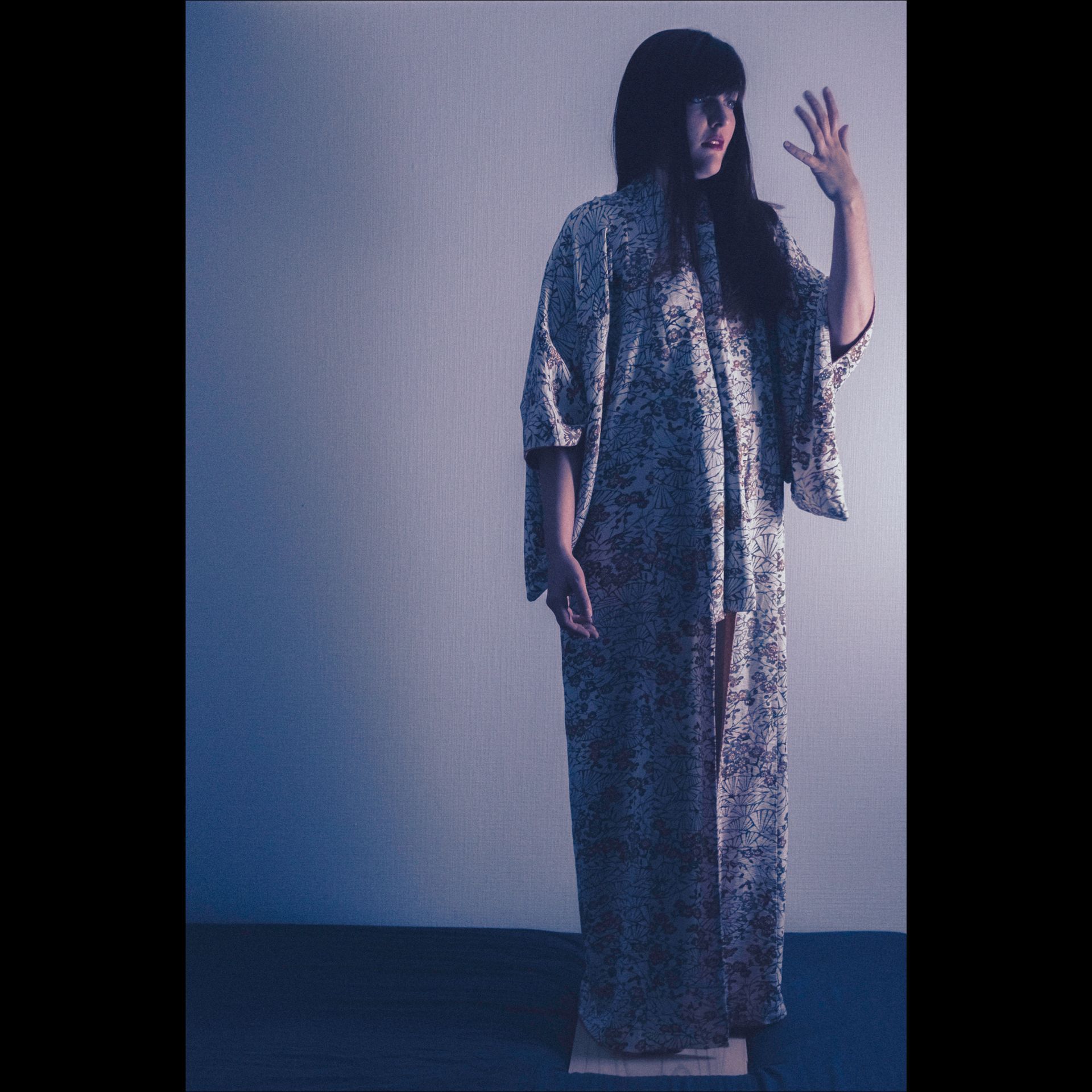 Woman in patterned dress raises hand, standing on a wooden plank against a blue-toned wall.