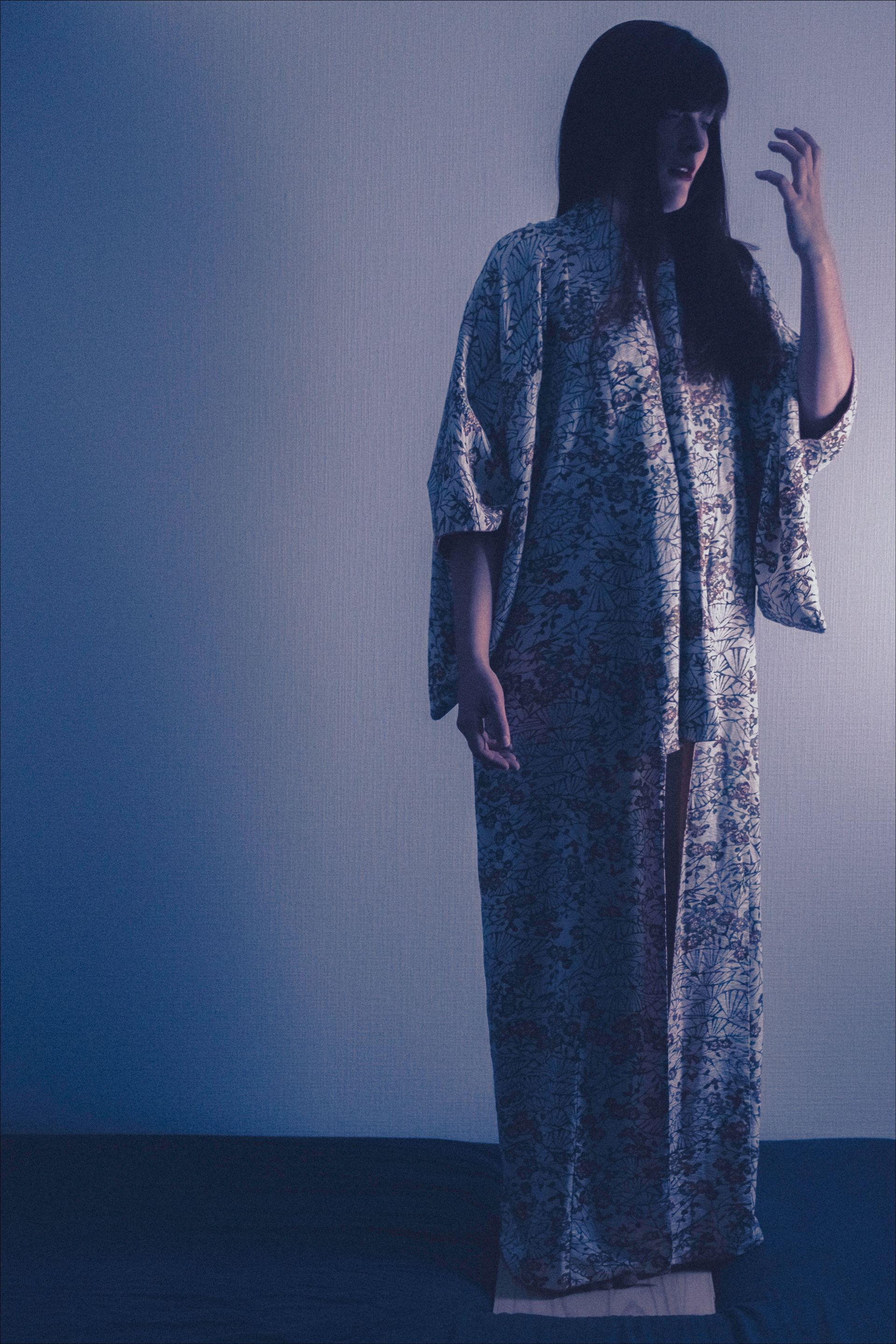 Woman in patterned kimono, gesturing with hand raised. Against a blue-lit wall, standing on a wooden surface.