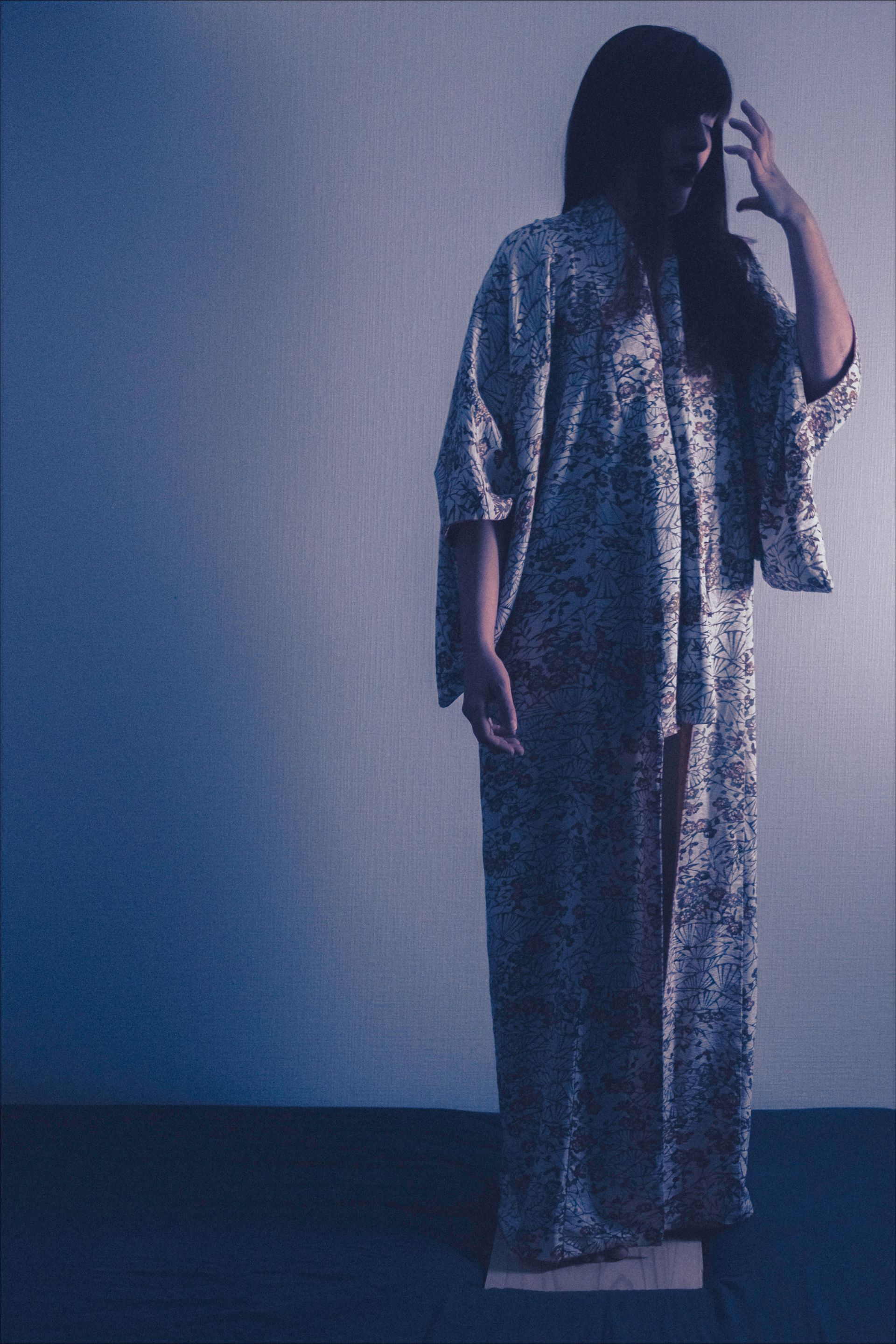 Person in patterned robe, standing, hand raised to head, against a plain blue-lit wall.