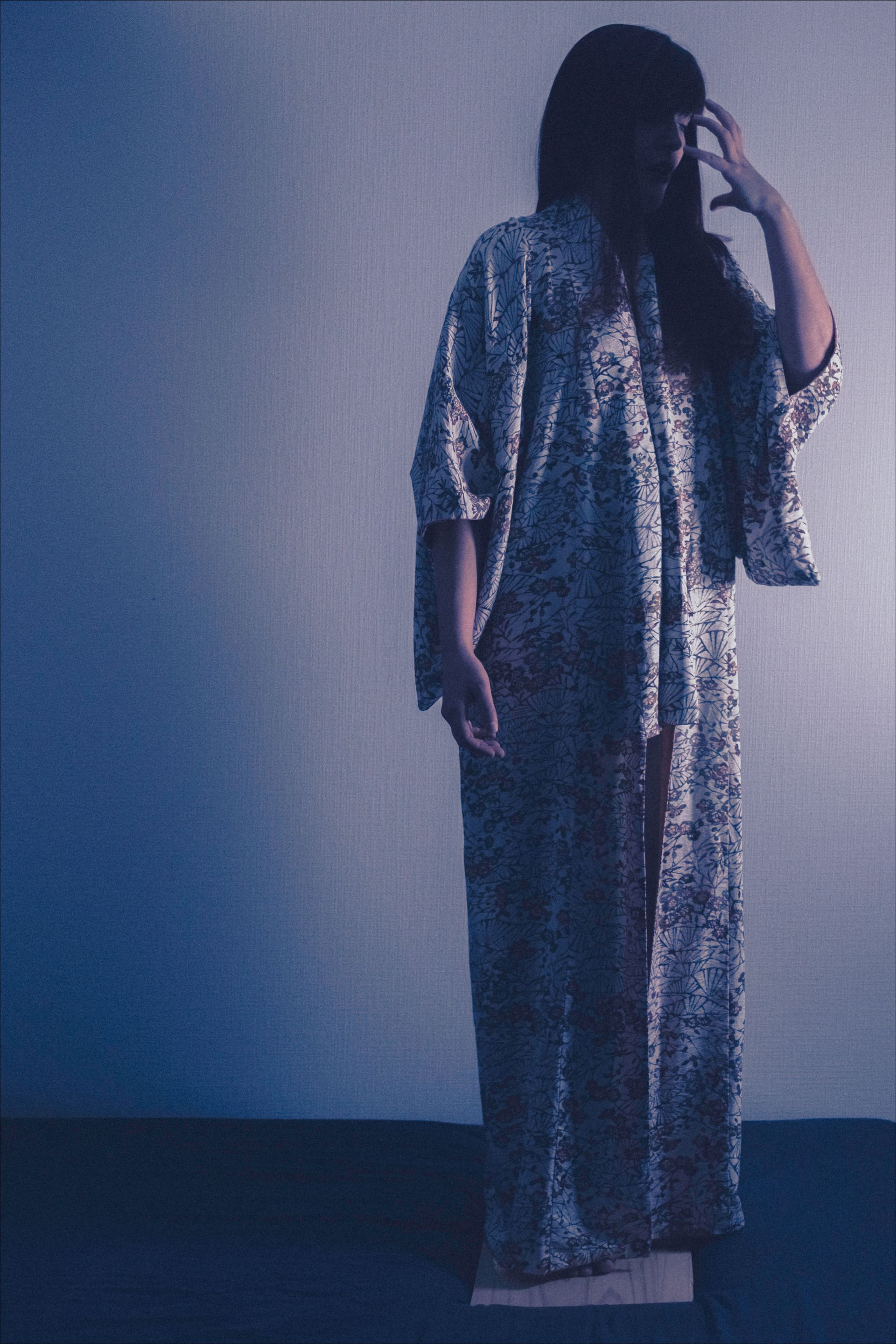 Person wearing a patterned robe, hands on head, standing in a dim space against a textured wall.