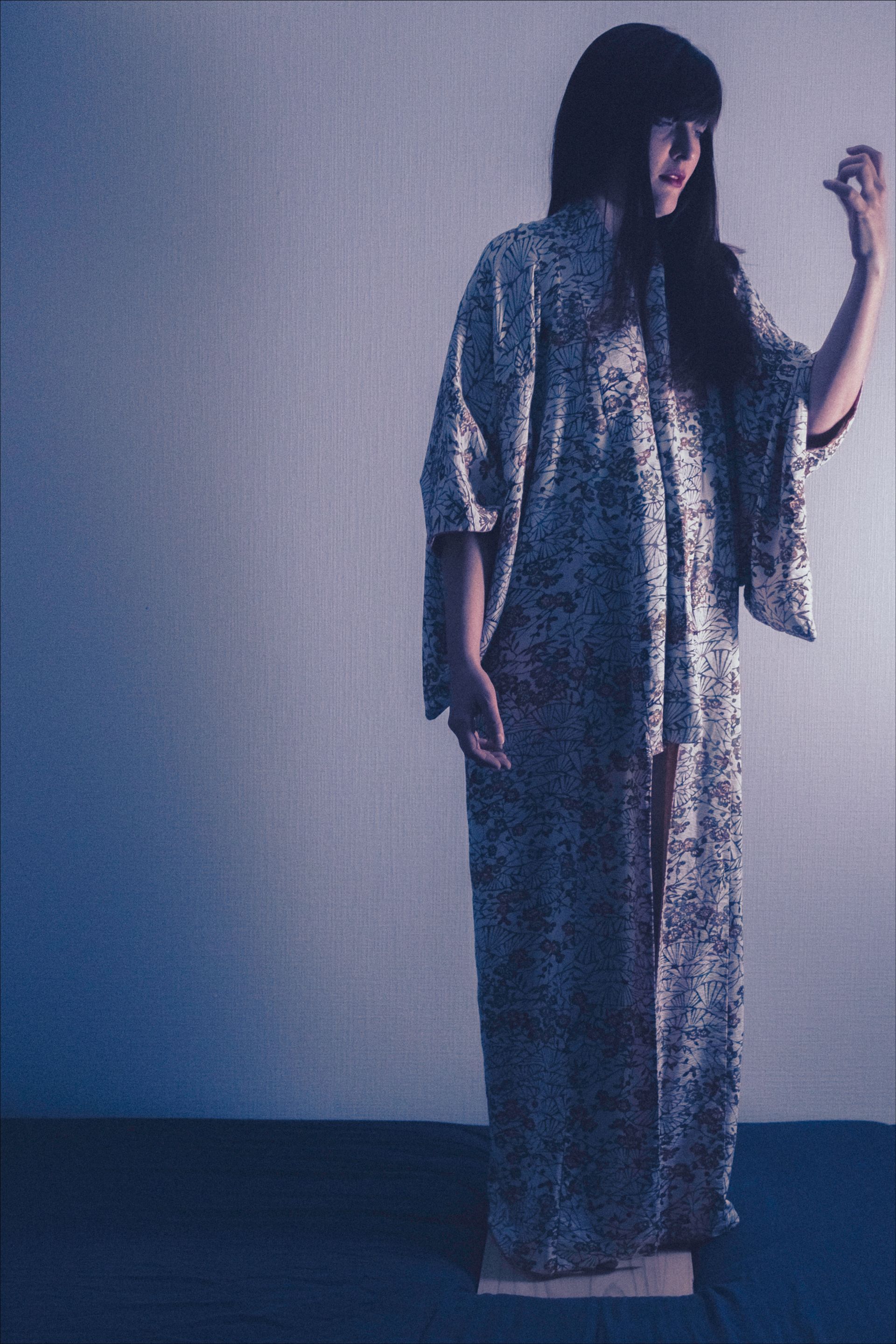 Woman in patterned robe, dark hair, raised hand, standing in front of a blue-toned wall.