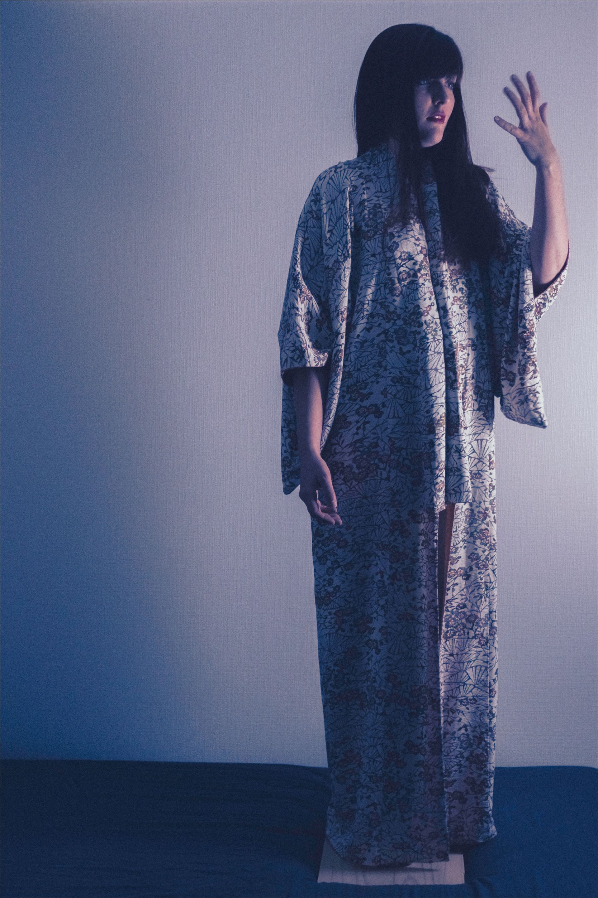 Person in patterned kimono raises hand, standing on a wooden platform against a blue wall.