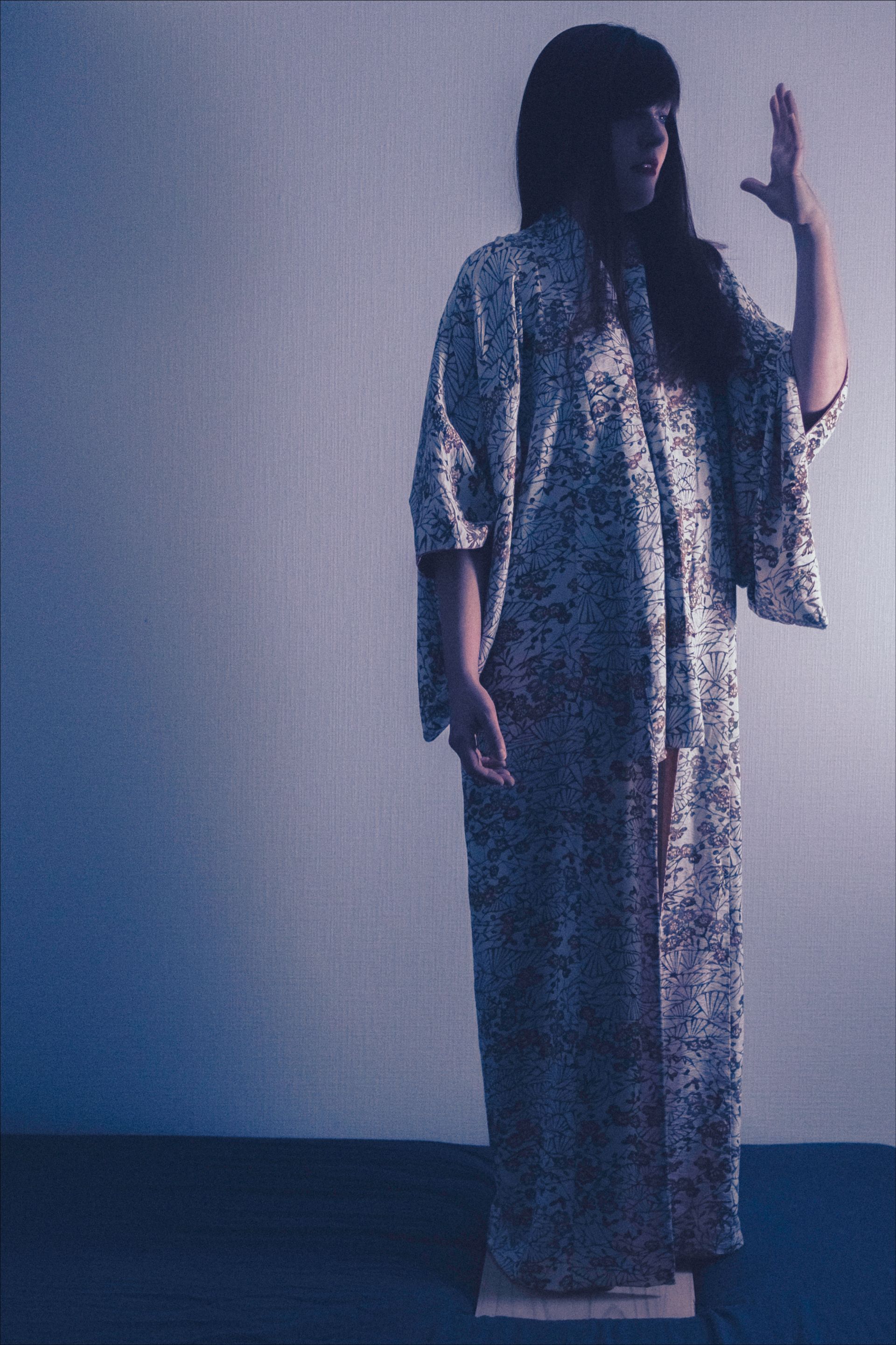 Woman in long patterned robe, hand raised, standing on platform in front of a white wall.