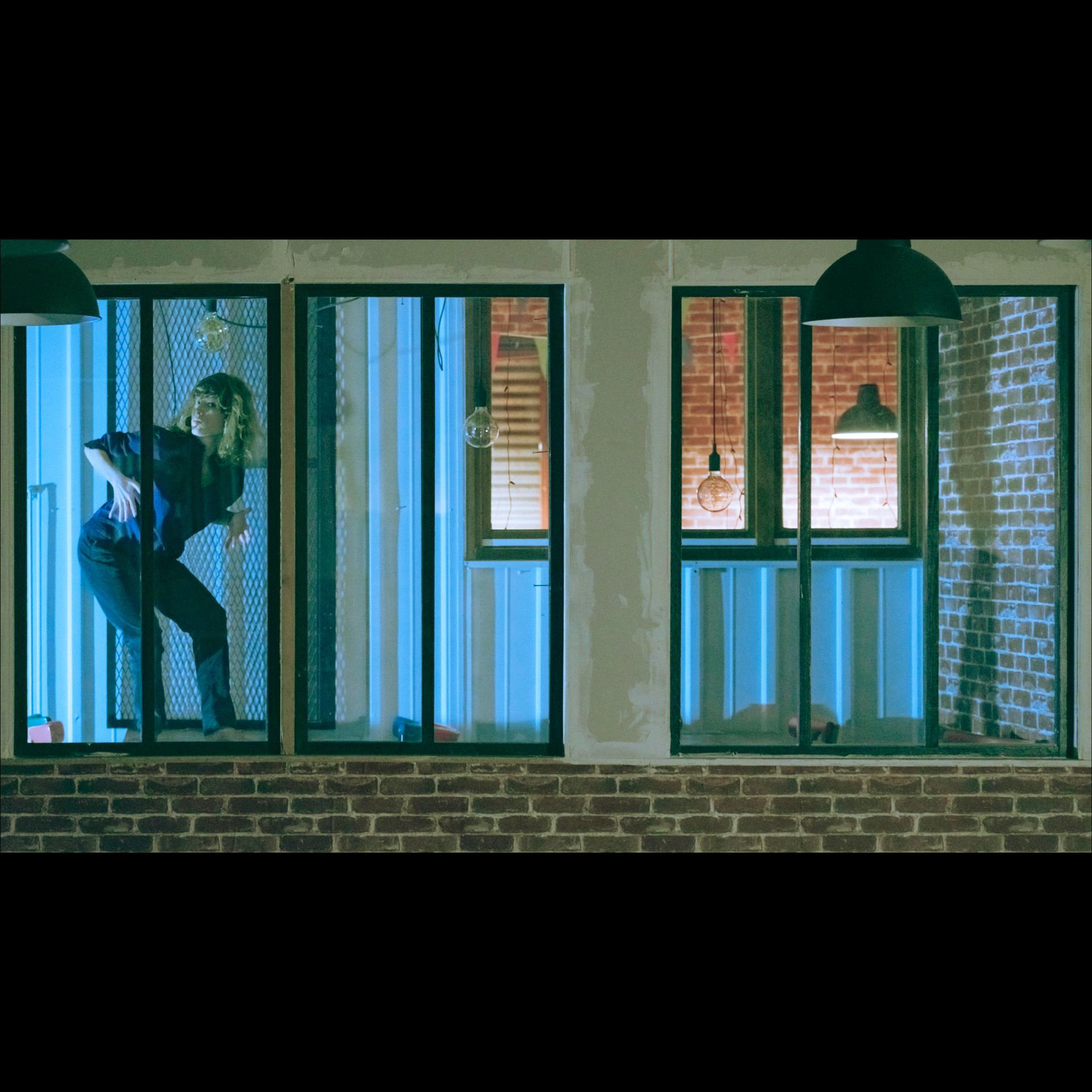 Person in a dark room illuminated by blue light, looking out a window. Brick exterior with pendant lights.