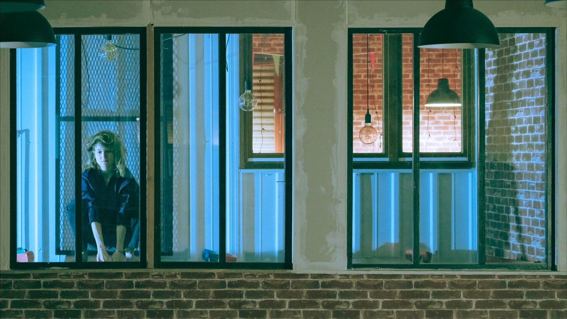 Woman in dark shirt leans on window. Windows frame industrial brick building, with contrasting blue and orange lighting.