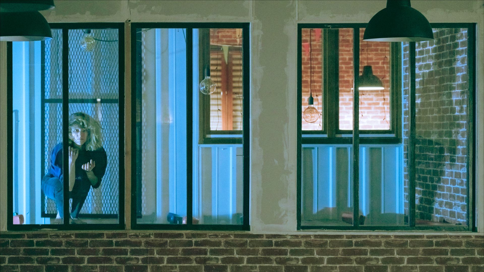 Woman peering through a window. Blue and orange lighting. Brick building exterior.