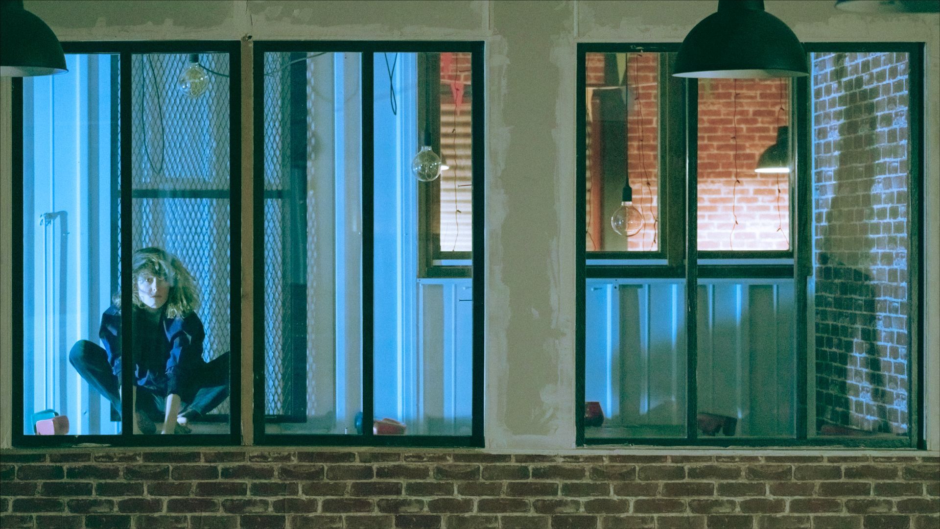 A person kneels near a window looking at a phone, lit with blue light. Night scene with brick wall and lights.