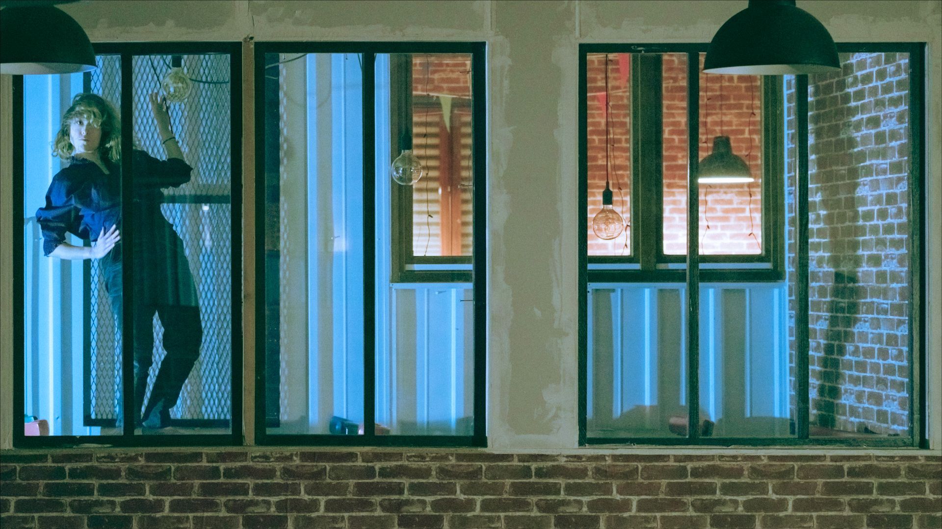 Woman inside a building with metal bars on the windows; blue and orange lighting.