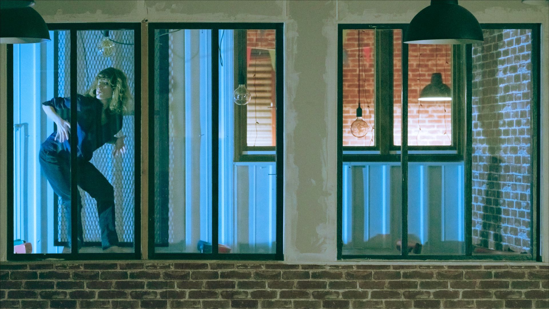 Woman peeking through a window with blue lighting. She is wearing a dark shirt and pants. Brick wall in the foreground.