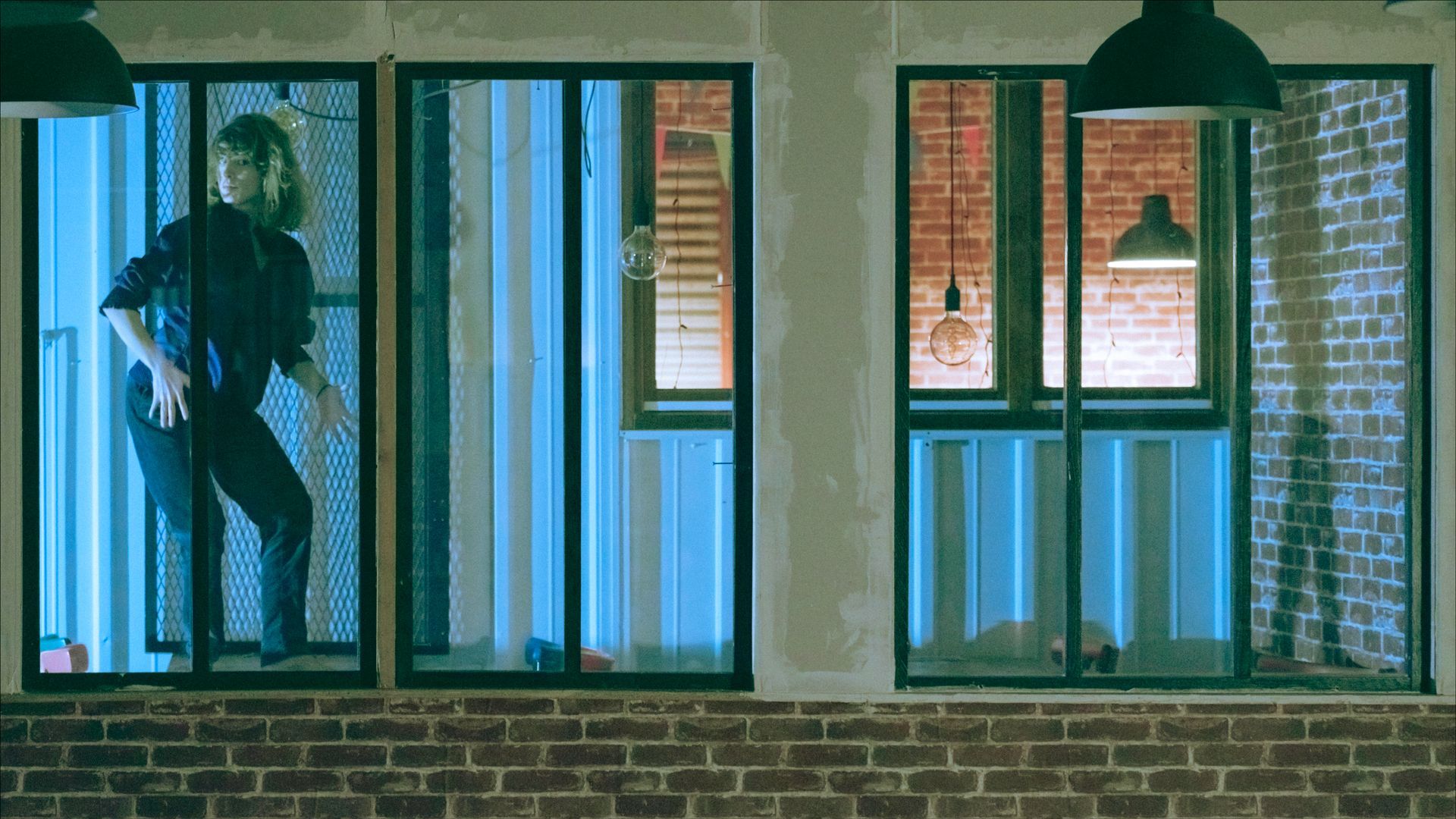 Person moving inside a room, viewed through window panes. Neon blue light. Brick building.