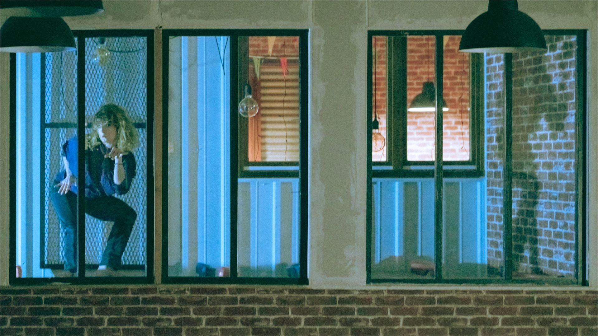 Woman peeking through a window with blue light, brick building exterior at night.