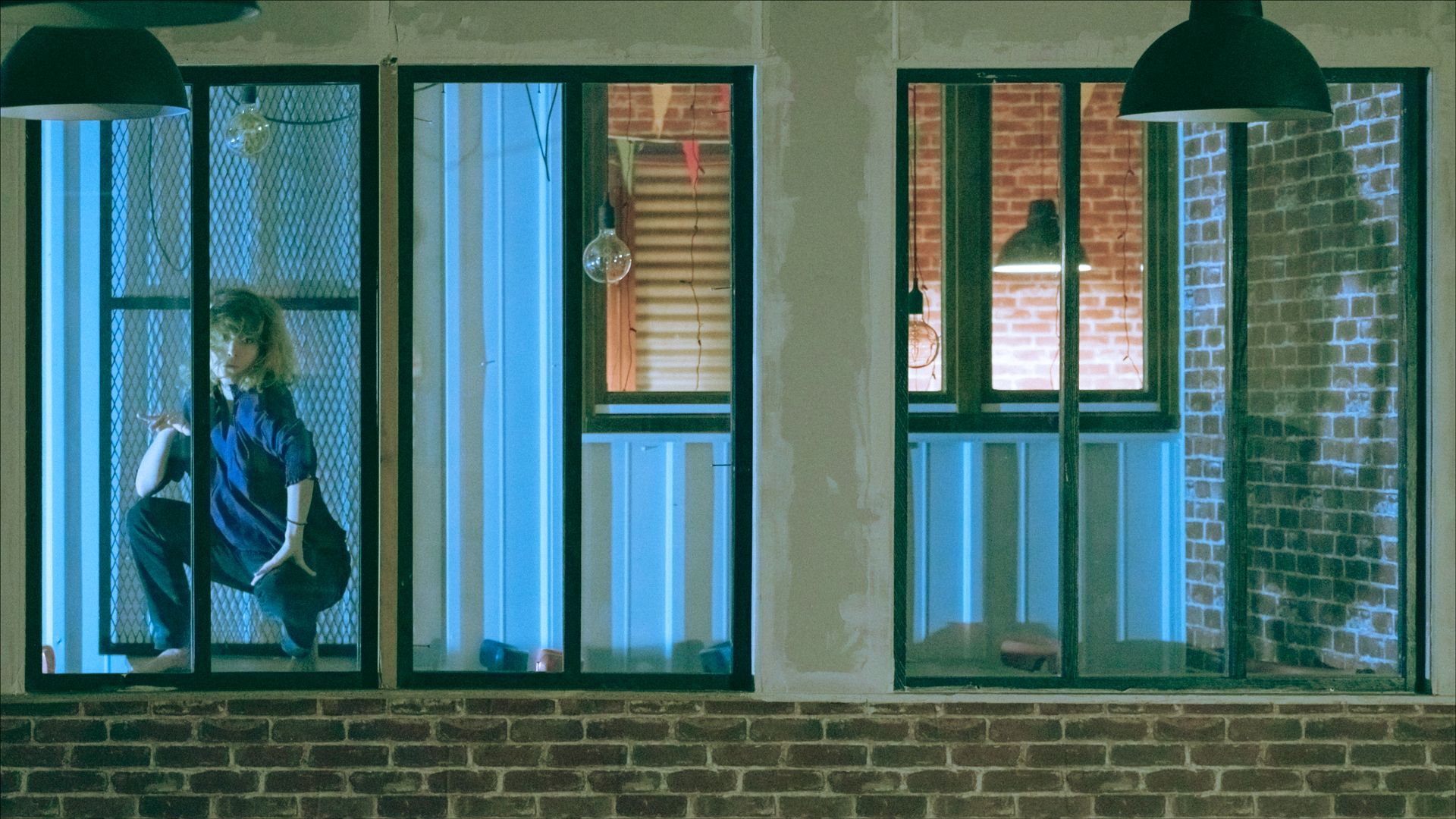 Person squatting by a window, looking outside. Nighttime scene with brick wall and blue-toned lighting.