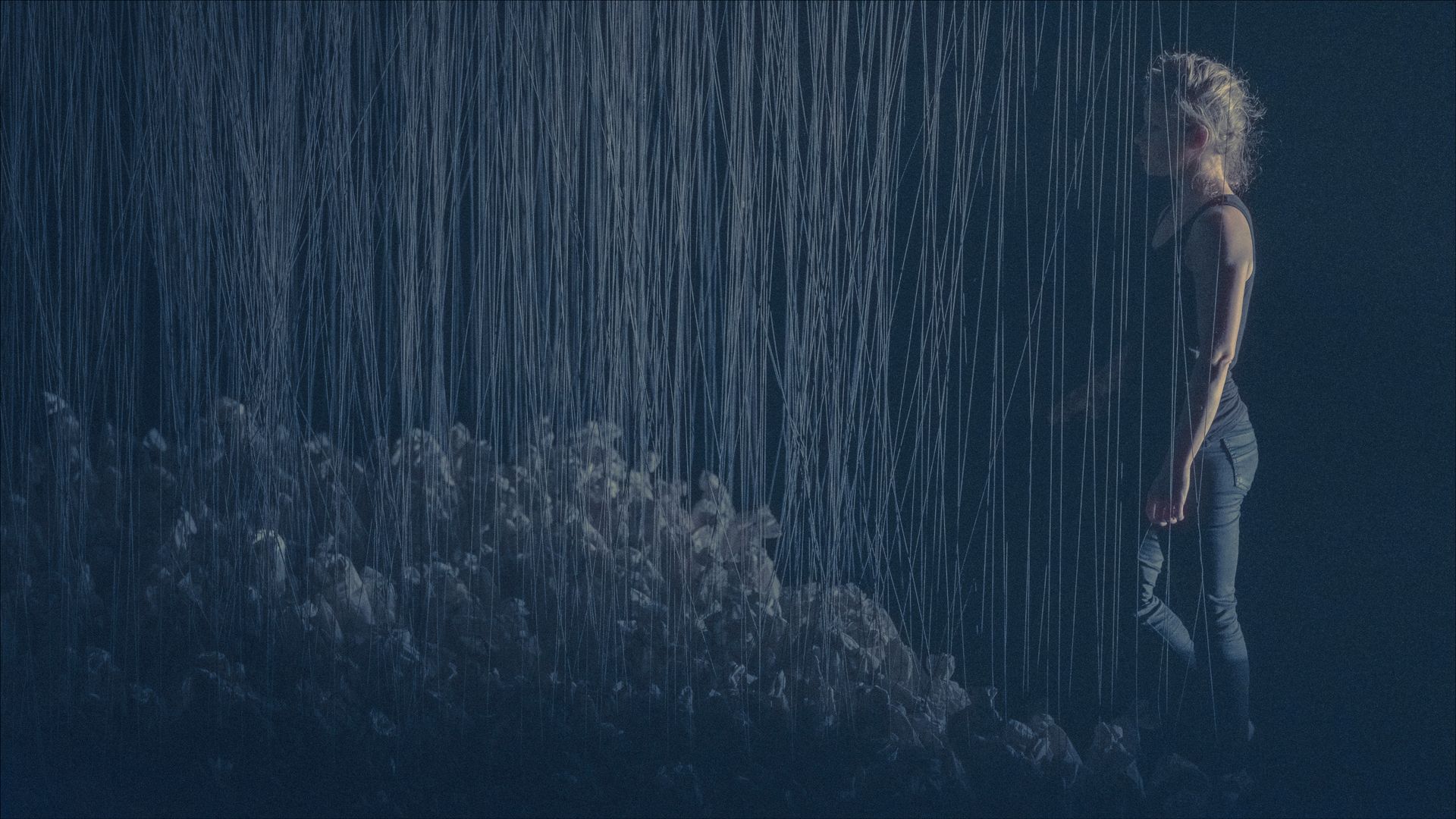 Woman standing in a dark space, partly obscured by a simulated rainfall cascading over a light-colored ground.