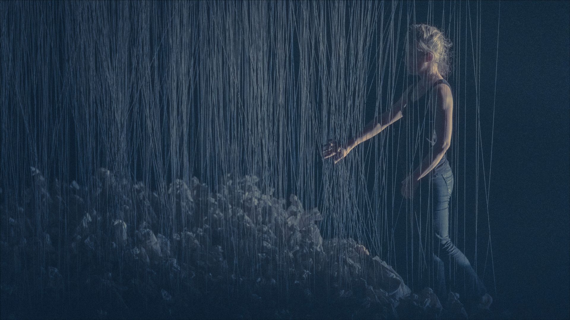 Woman in jeans and top, reaching out through simulated rainfall.