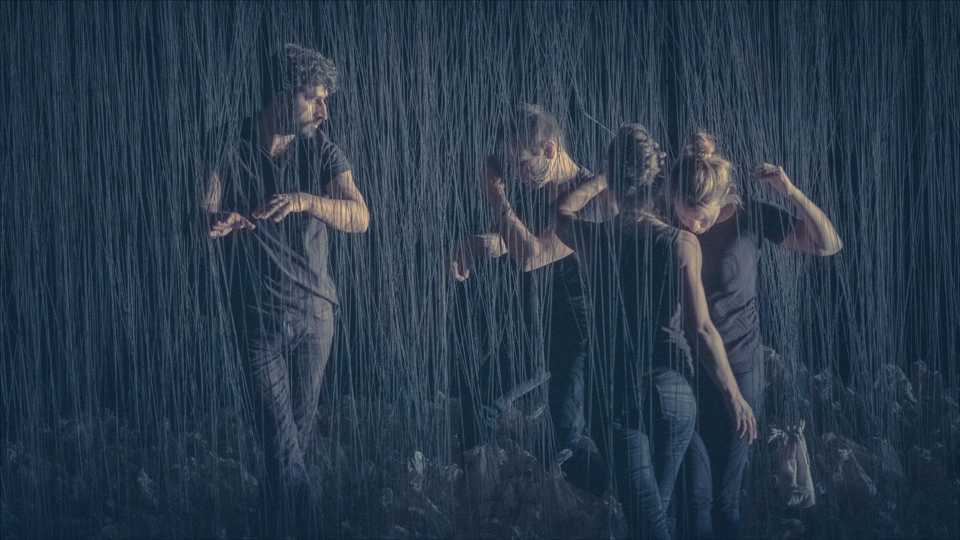 Person standing in rain, multiple figures blurred dancing. Dark tones, vertical rain lines.