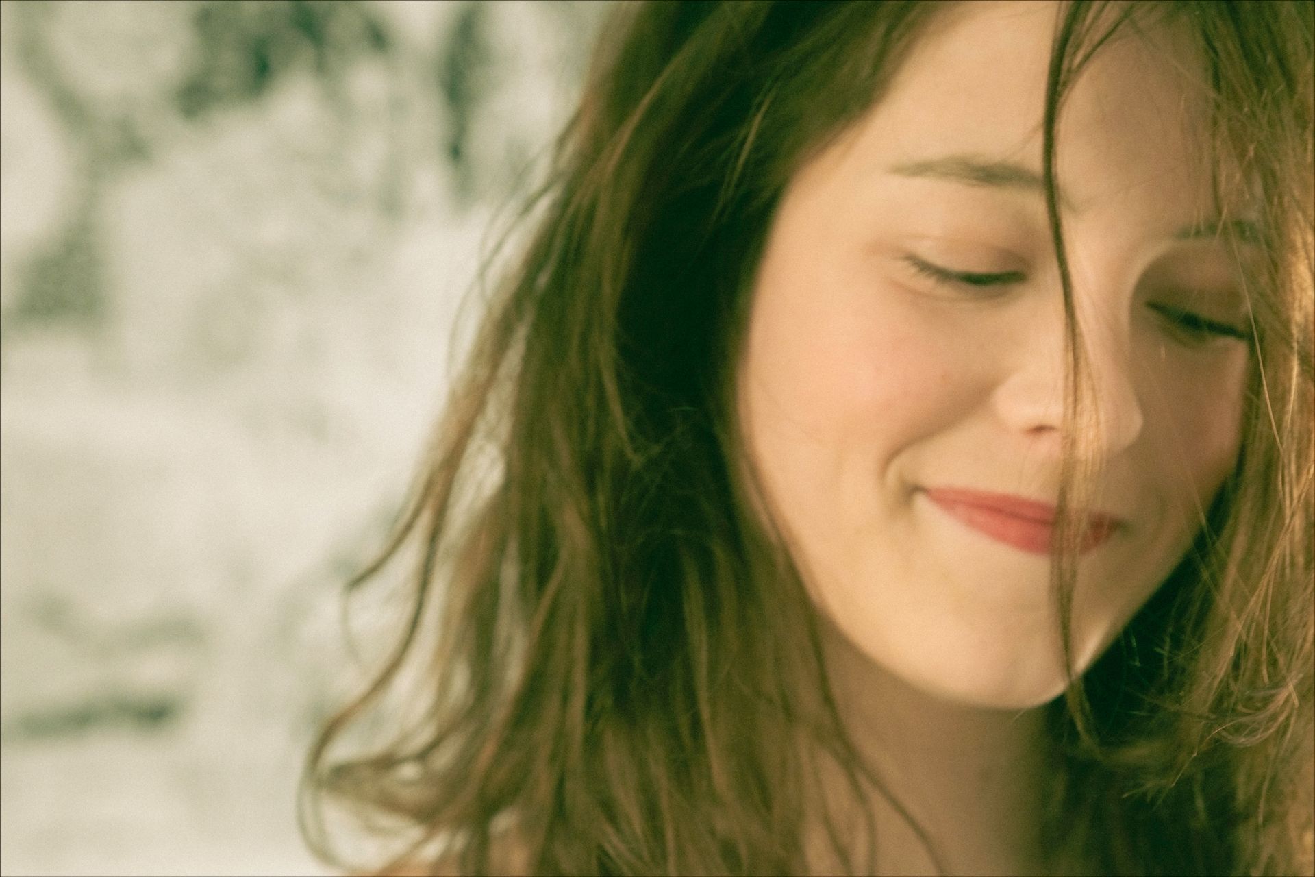 Woman with closed eyes, smiling softly, with loose brown hair; blurry background.