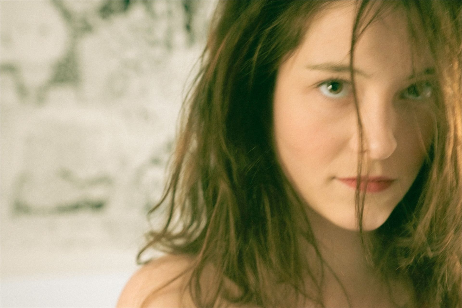 Woman with light skin and messy brown hair looks at the viewer with a neutral expression; background is a blurred drawing.