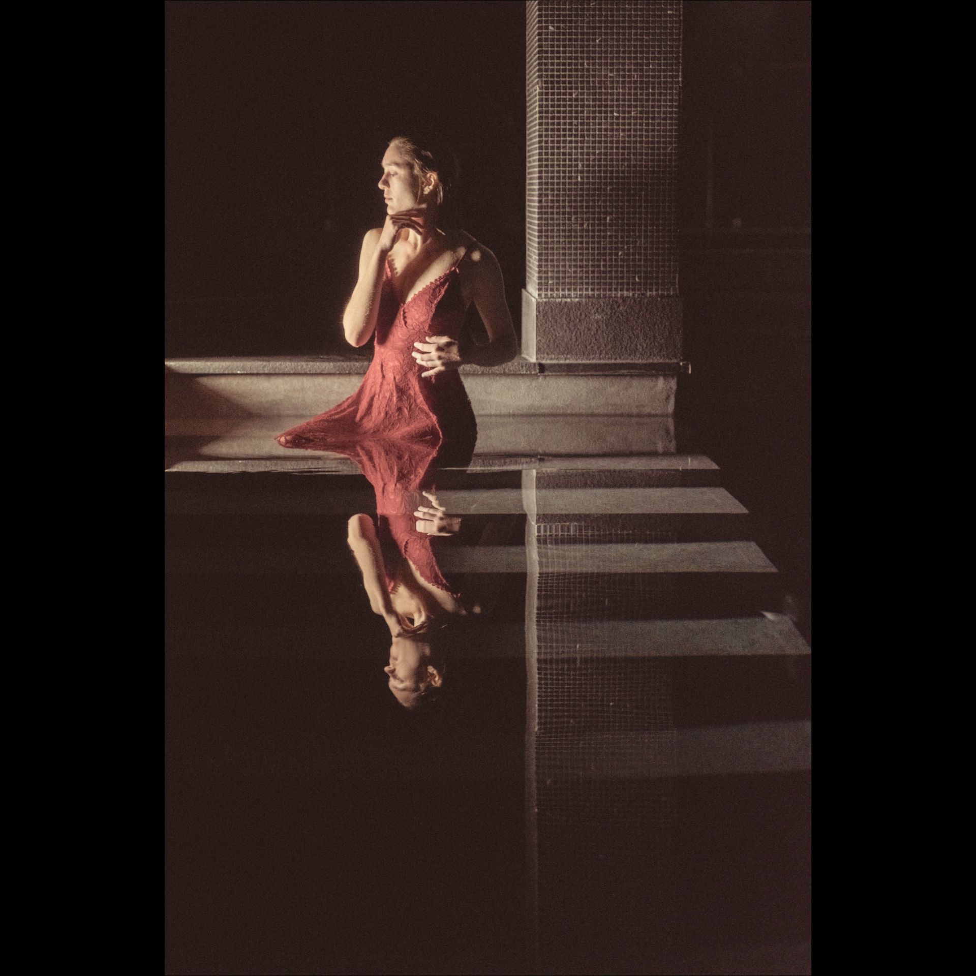 Woman in red dress, reflected in water, stands by tile steps; dark background.