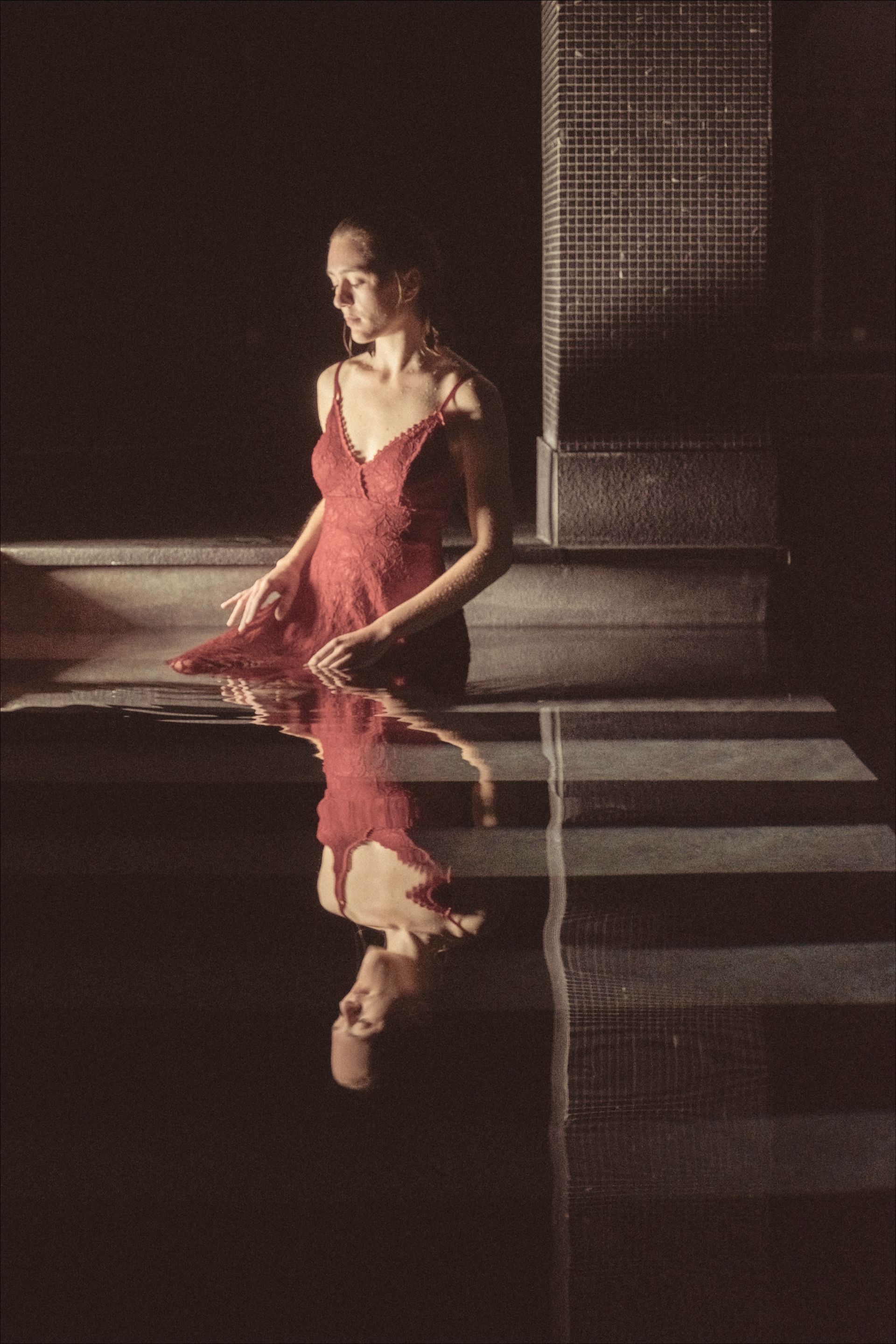 Woman in red dress sits by water, reflected image. Dark setting, soft light.