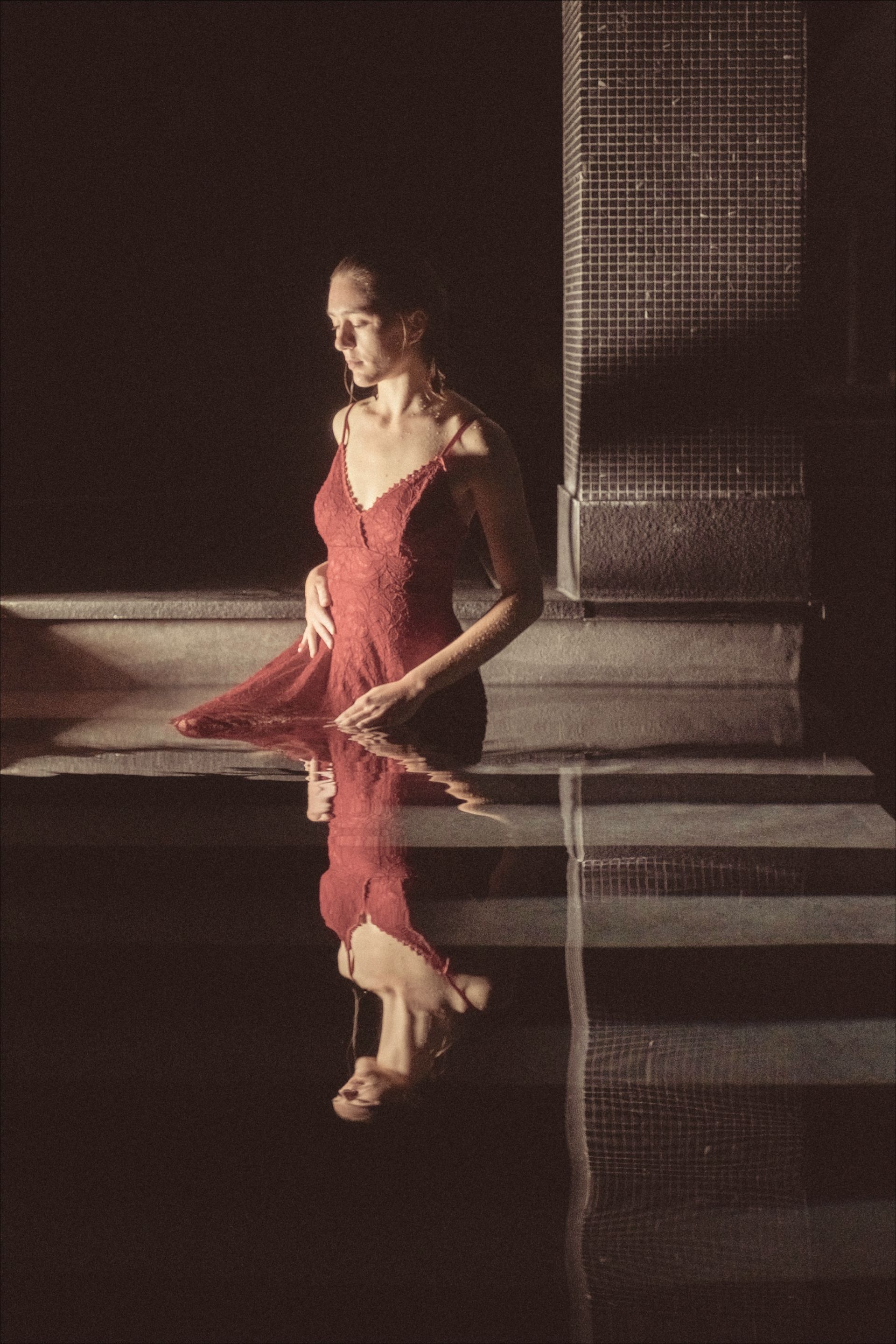 Woman in red dress standing in water, gazing downward. Water reflects her and nearby tiled wall. Dark setting.