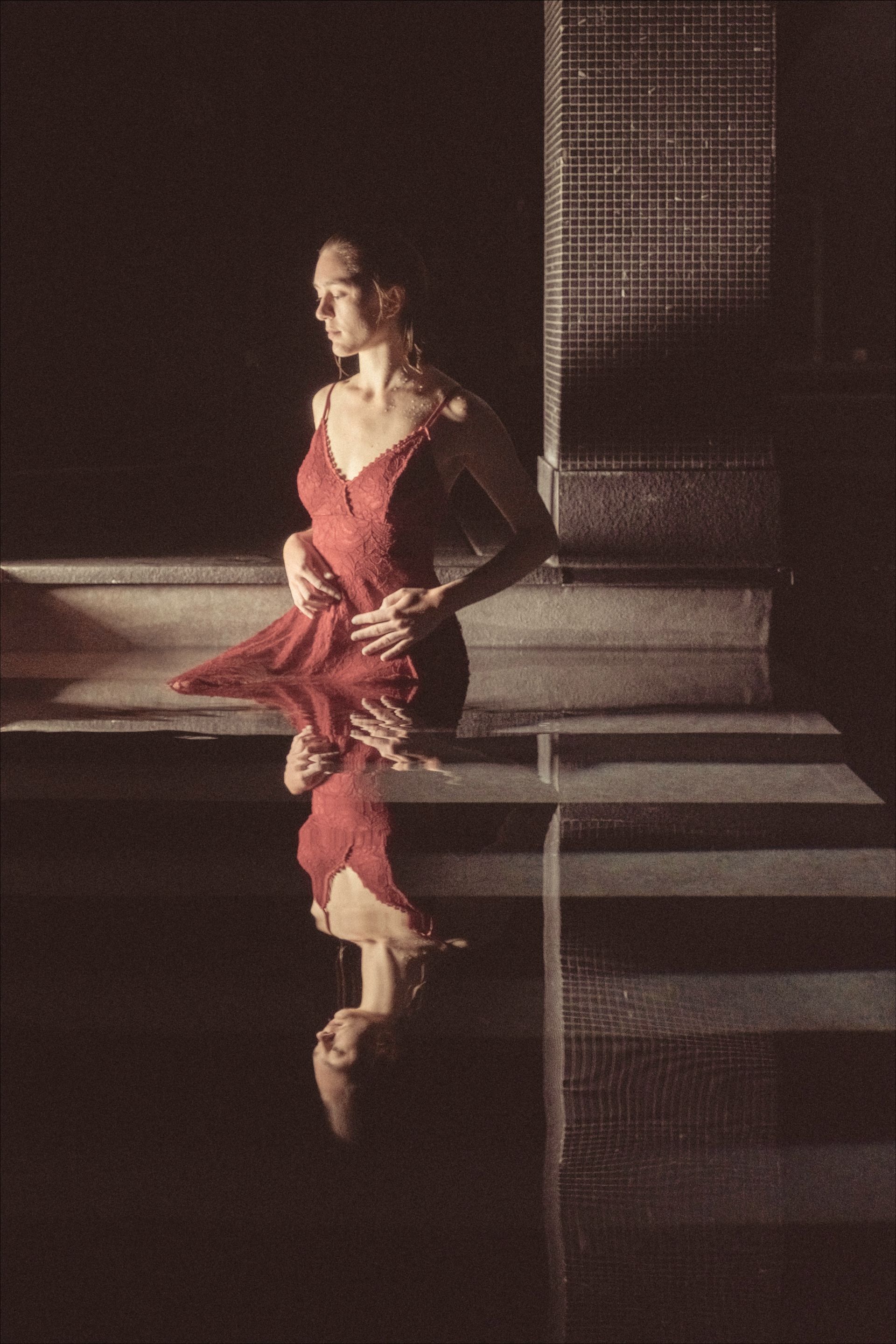 Woman in red dress, standing by water, reflected image. Dark setting, tiled wall.