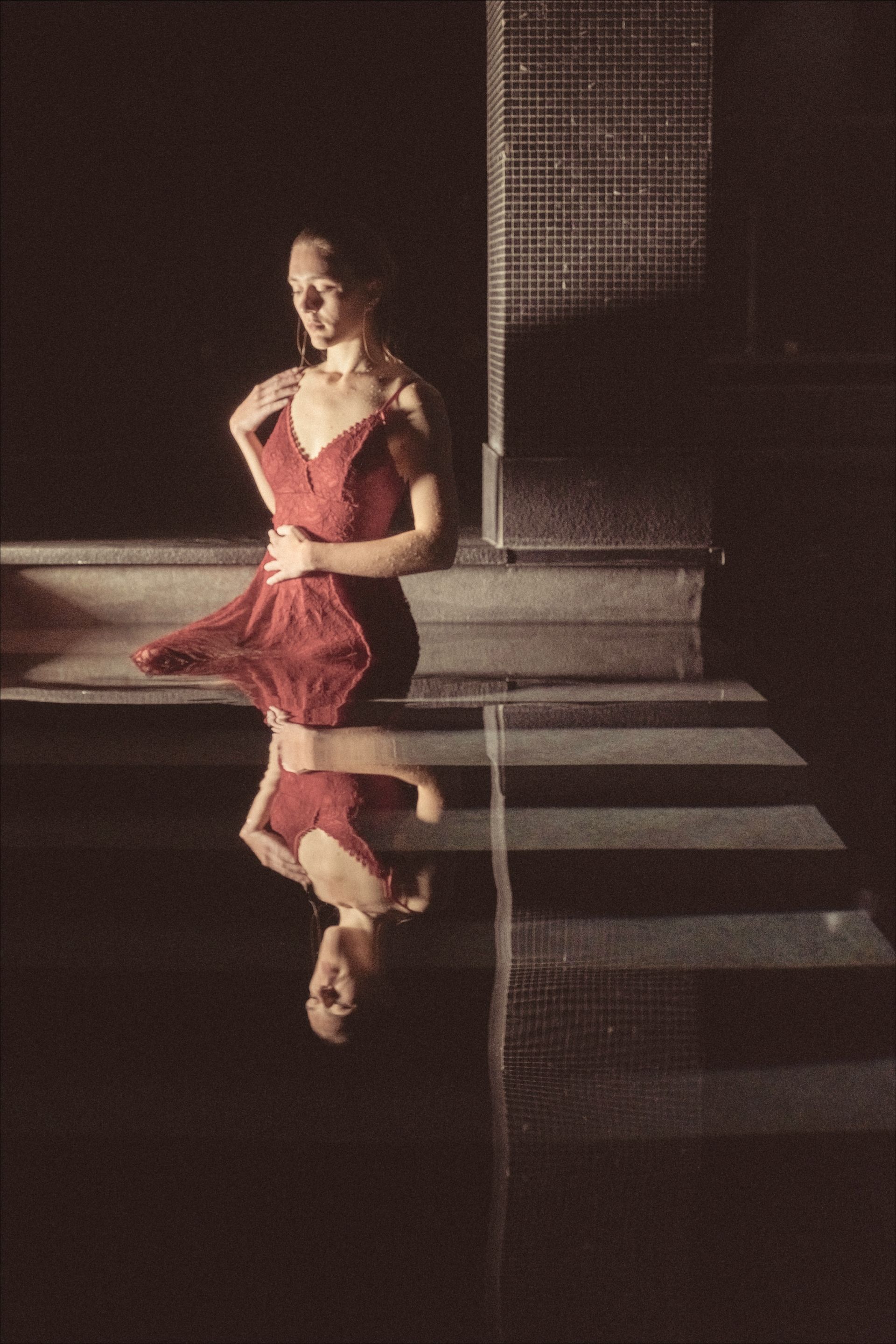 Woman in red dress reflected in water, standing on steps in a dark, architectural setting.