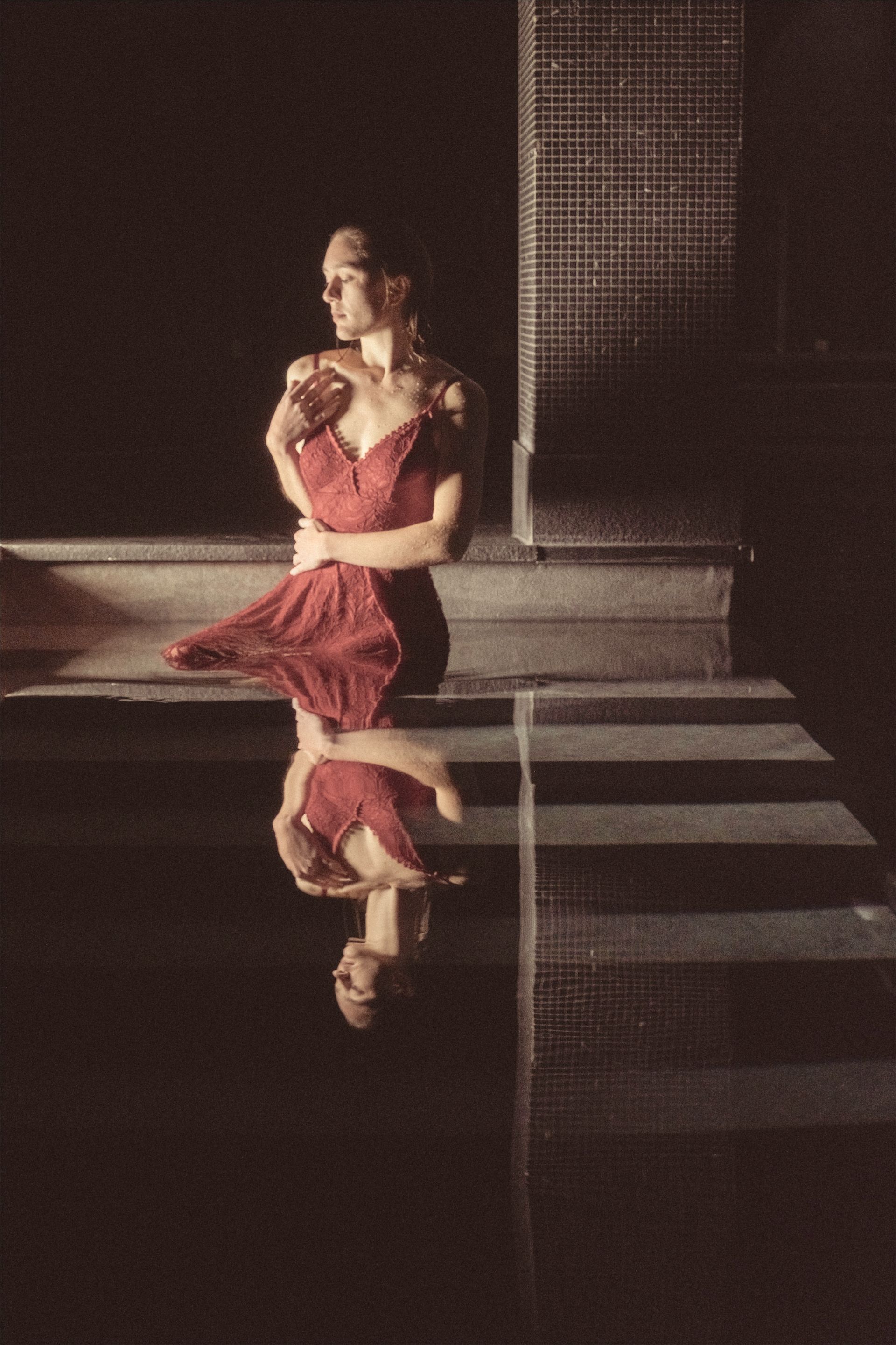 Woman in red dress standing near water, reflection visible. Dark setting with tiled wall and stairs.