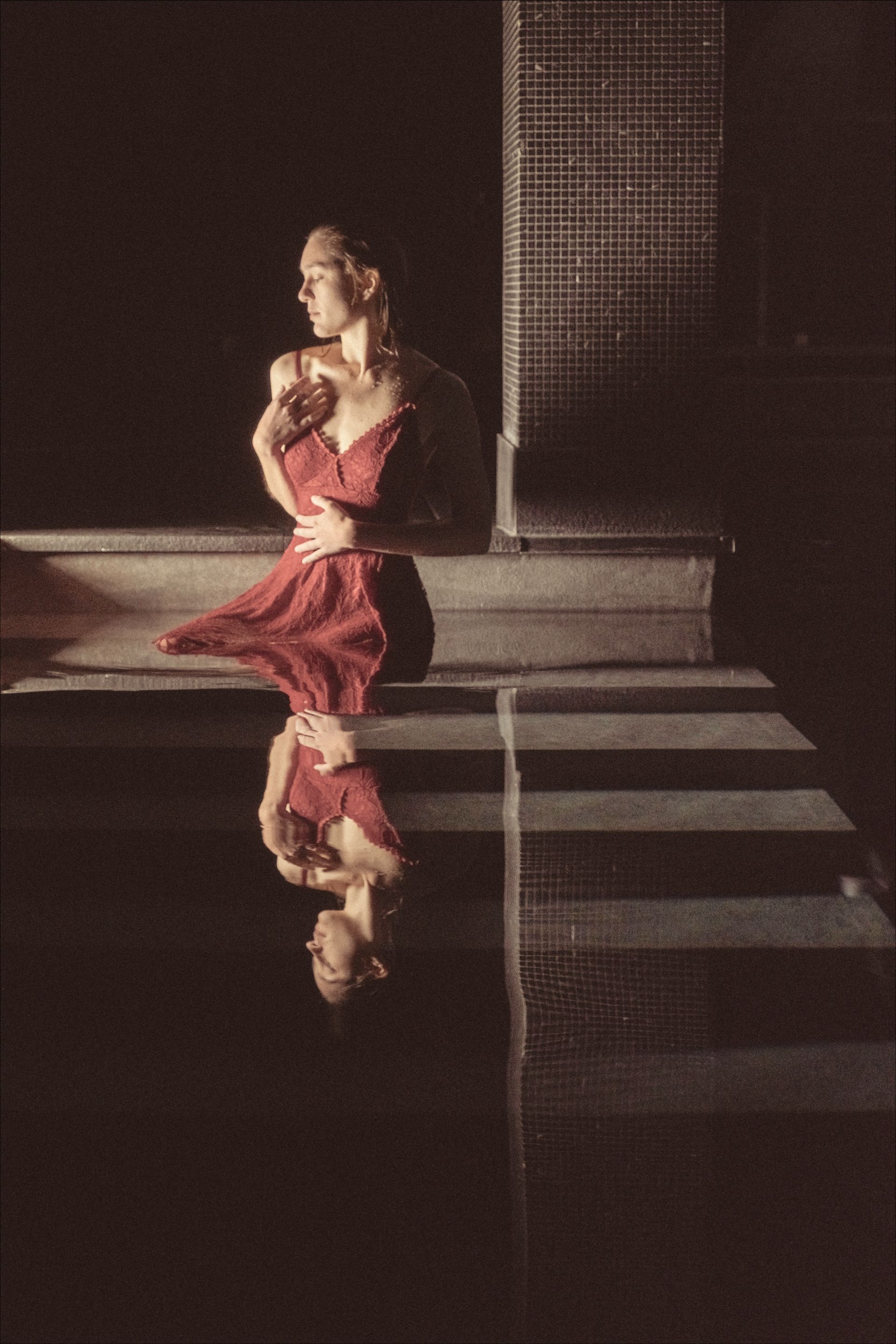 Woman in red dress, reflected in water, stands by brick structure.